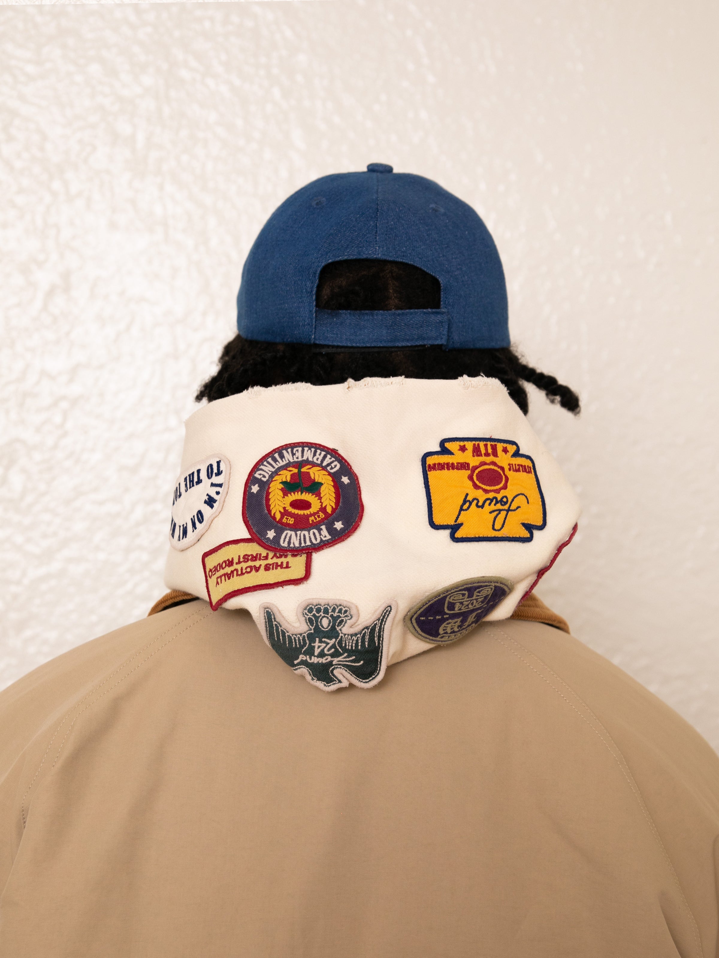 A person in a vintage look, wearing a beige jacket and blue cap, stands with their back to the camera. Draped over their shoulders is a cloth similar to FOUND's Faded Patch Hoodie, featuring colorful patches that evoke nostalgic charm.