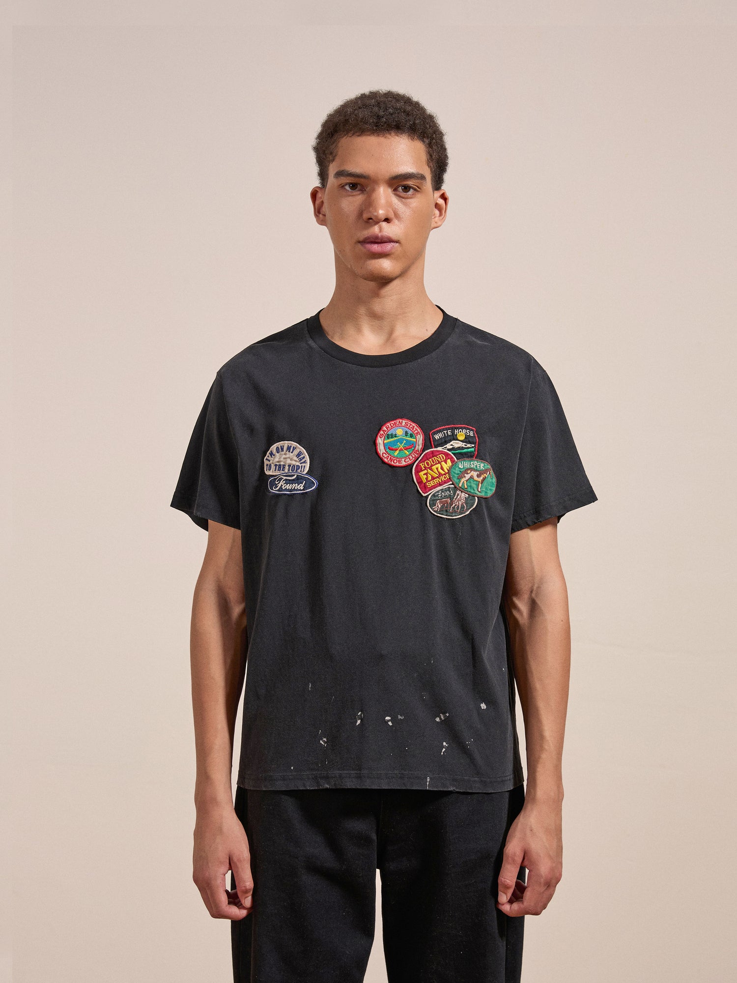 An individual with short curly hair is wearing the Multi Patch Washed Tee by FOUND and black pants, standing against a plain background.