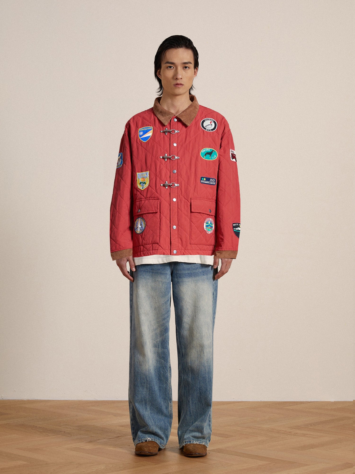 A person with long hair stands facing forward, wearing a red Farmstead Quilt Patch Jacket by Found and wide-leg blue jeans, against a plain indoor backdrop.