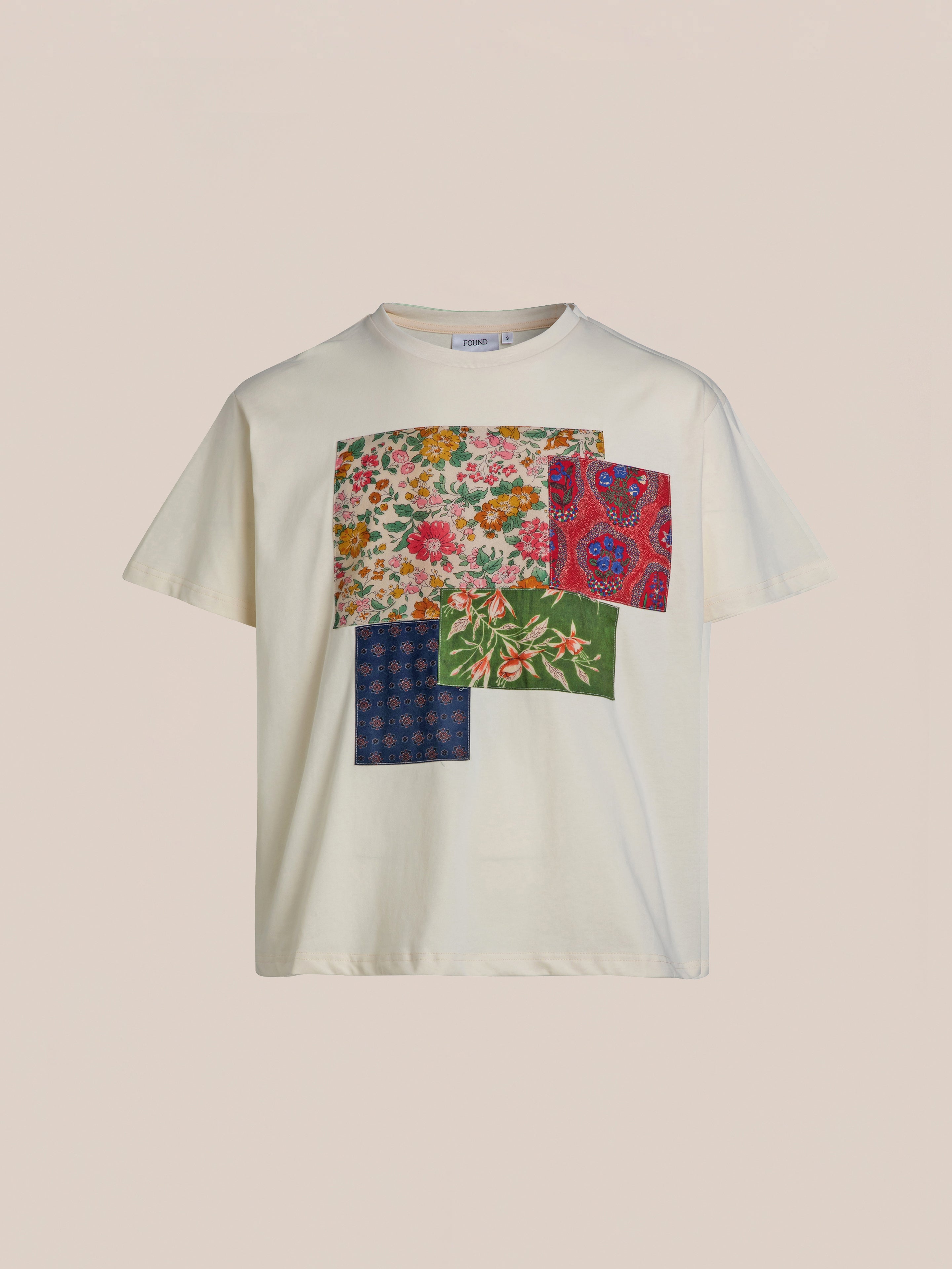 The Patchwork Tee by FOUND is a white short-sleeve T-shirt featuring a vibrant patchwork design with floral and paisley patterns in various colors on the front.