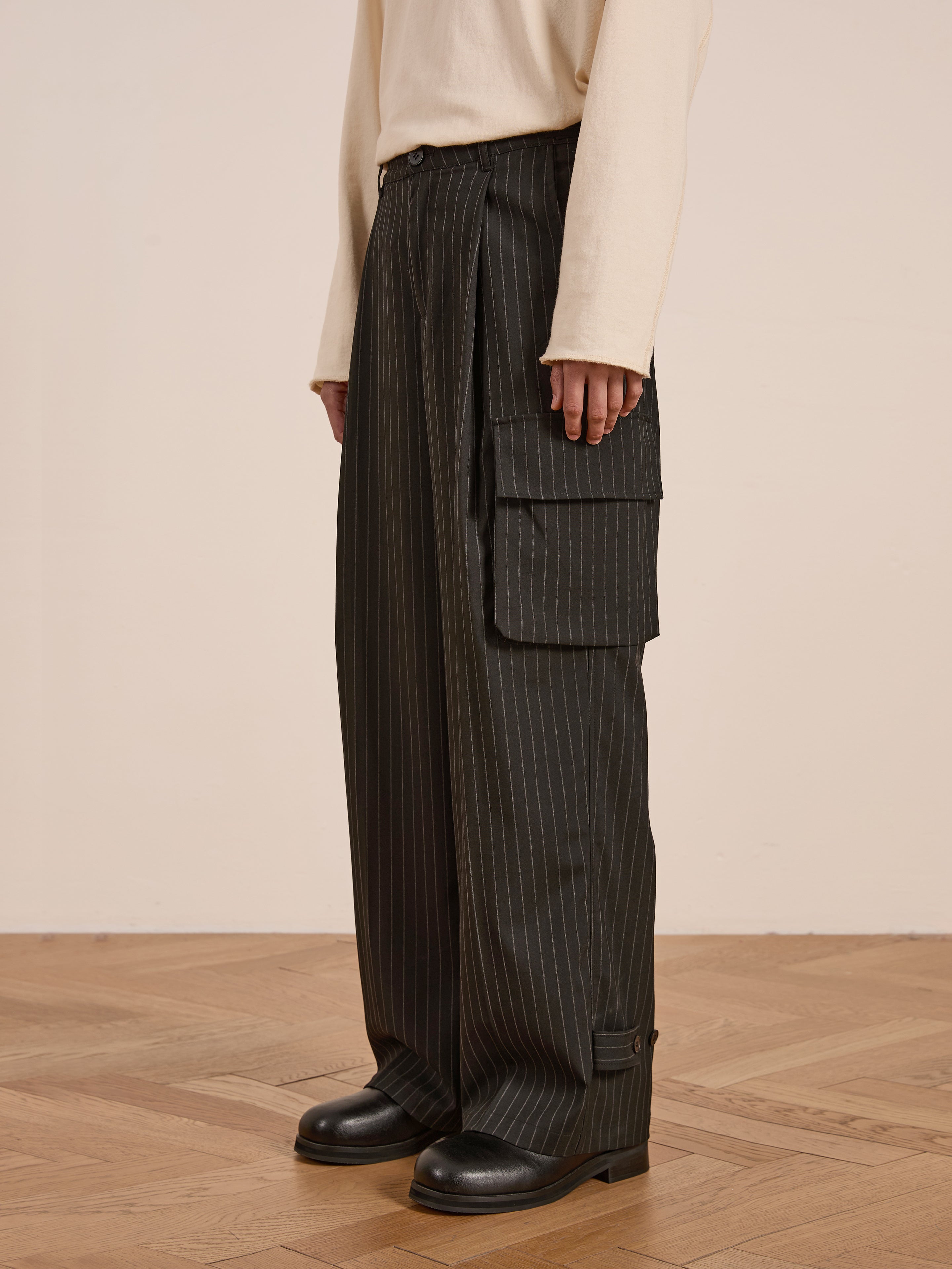 A person stands on a wooden floor wearing FOUND's Hava Pinstripe Pleated Cargo Trousers in black, crafted from cotton-blend fabric, paired with a cream long-sleeve shirt and sleek black shoes.