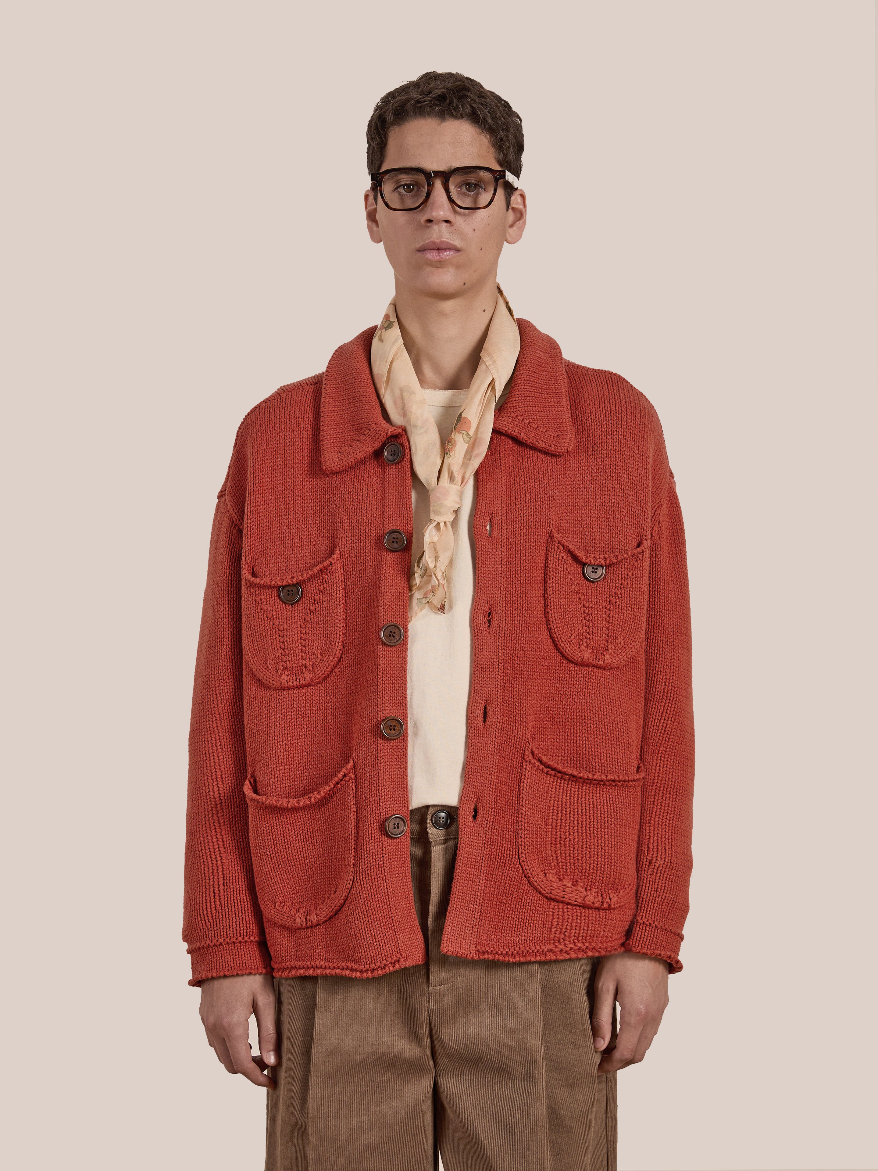 A person sporting glasses, a red Hunter Knit Collared Cardigan by FOUND, a beige scarf, and brown pants stands against a plain background. This chic ensemble elegantly highlights the art of transitional layering.