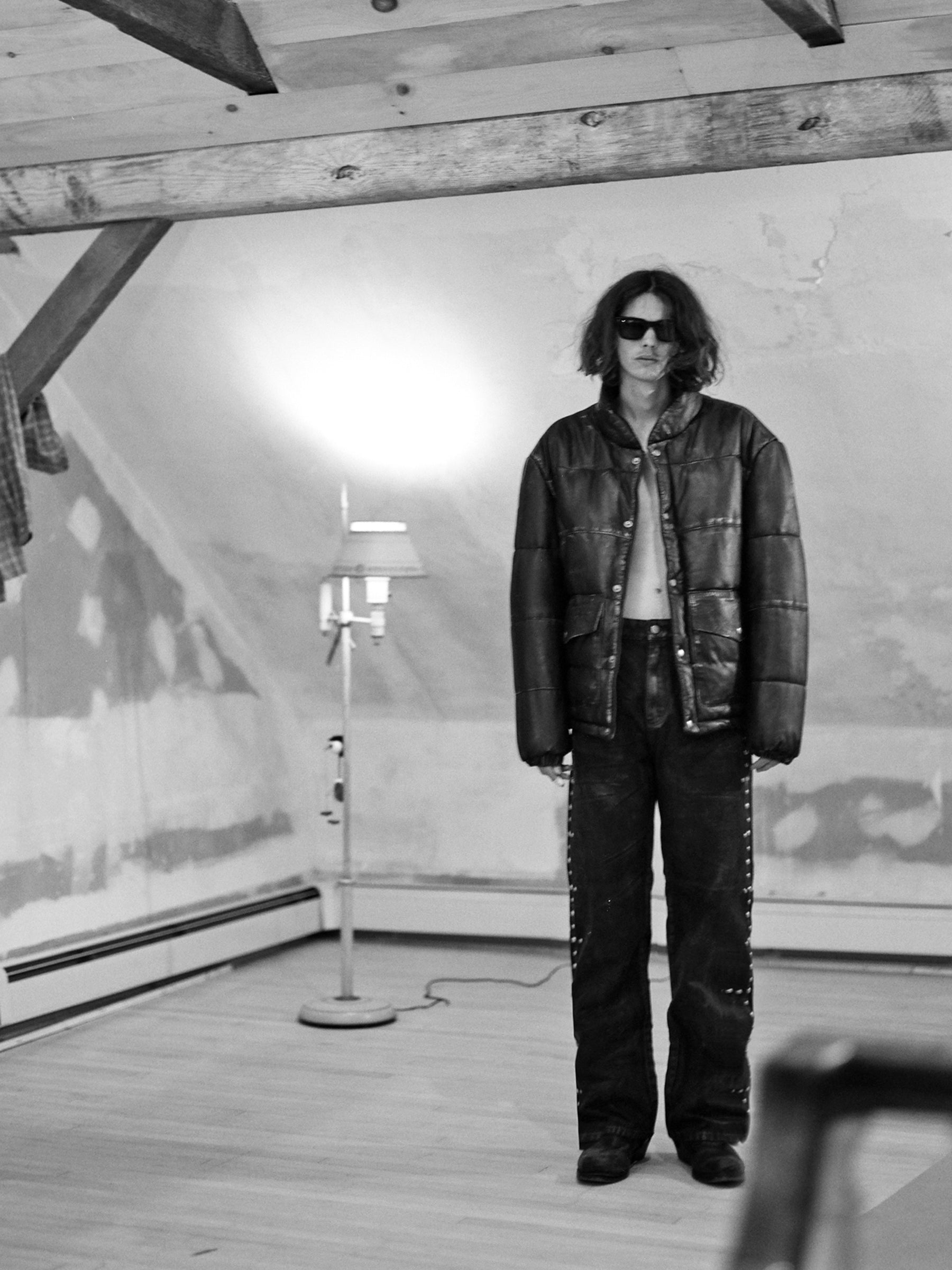 In a black and white attic scene, someone sports FOUND's Leather Distressed Puffer Jacket, stylish sunglasses, and studded jeans with a floor lamp in the backdrop.