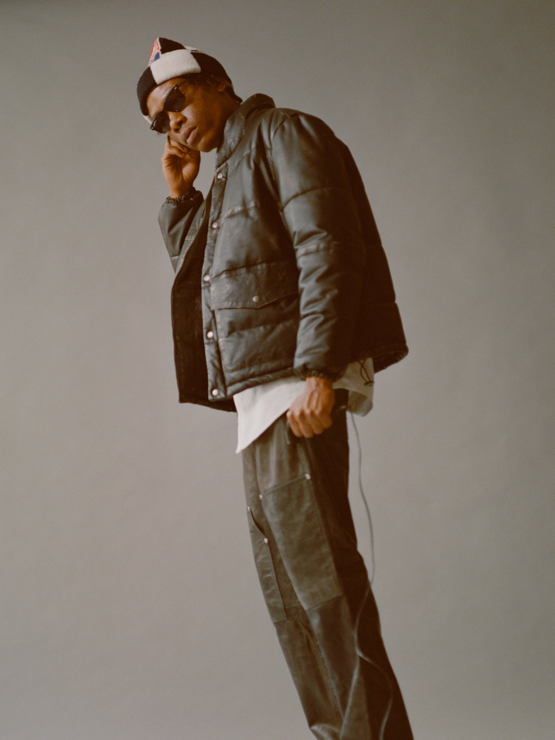 Someone, dressed in a Leather Distressed Puffer Jacket by FOUND, along with pants, a hat, and sunglasses, holds a phone to their ear against a plain background.