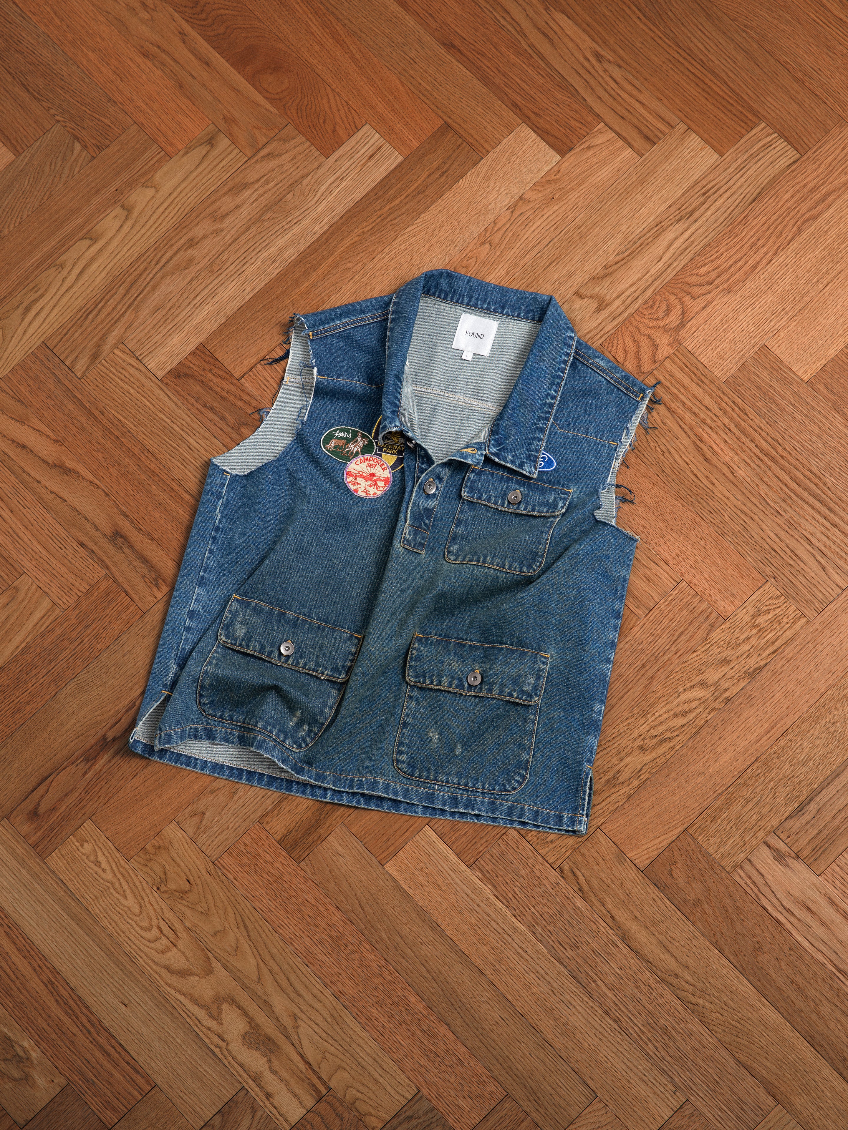 A vintage-inspired Raw Cut Patch Mechanic Denim Vest by Found with embroidered patches on a wooden floor.