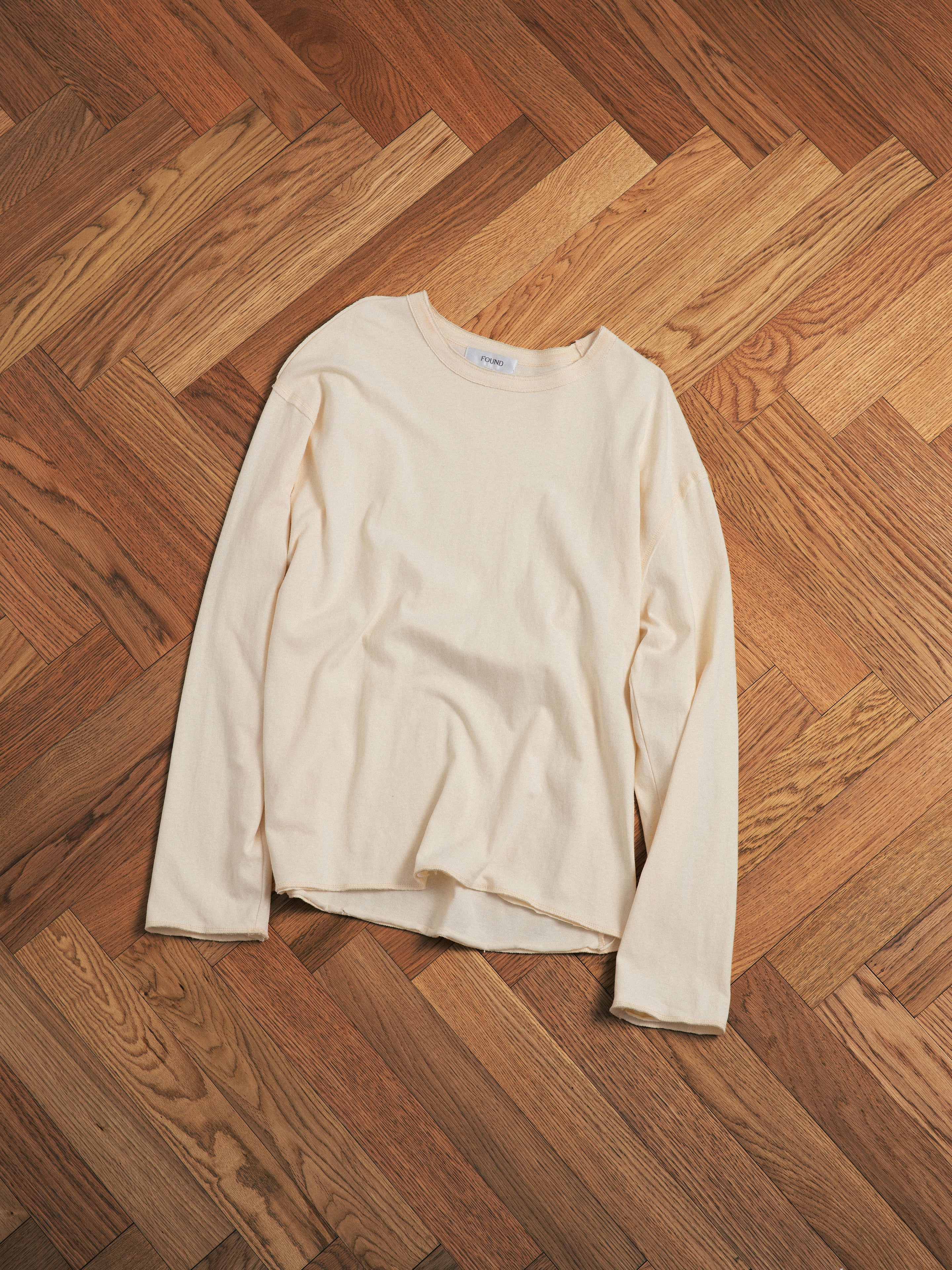 A white long-sleeved found Reversed LS Tee, crafted with premium materials, lies on a wooden floor.