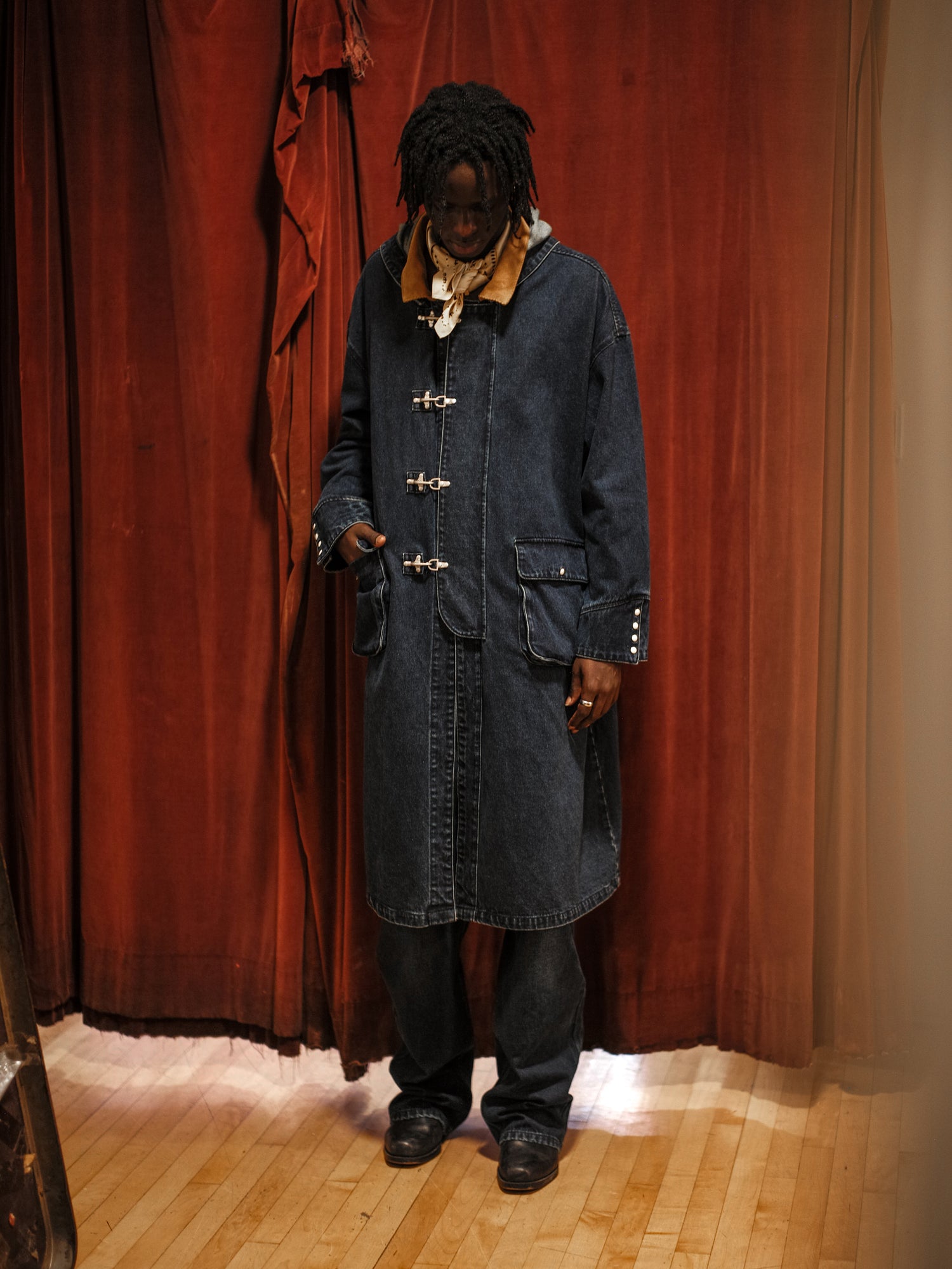 A man in a vintage fireman coat, the Found Sargasso Denim Buckle coat, standing in front of a red curtain.
