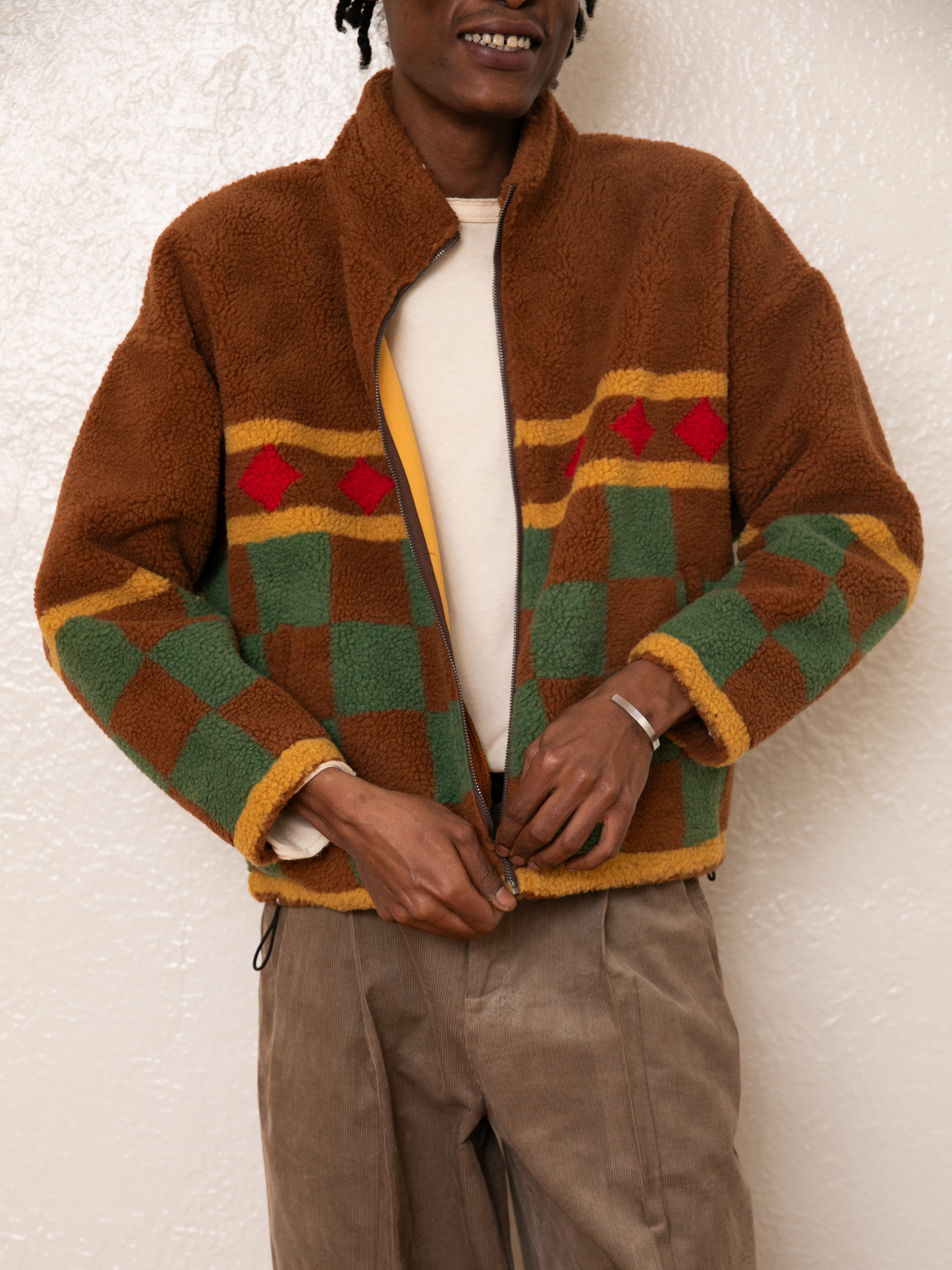 The person is zipping up a FOUND Checkered Zip-Up Sherpa jacket in brown, green, and red over a beige sweater, featuring an antique silver YKK zipper.