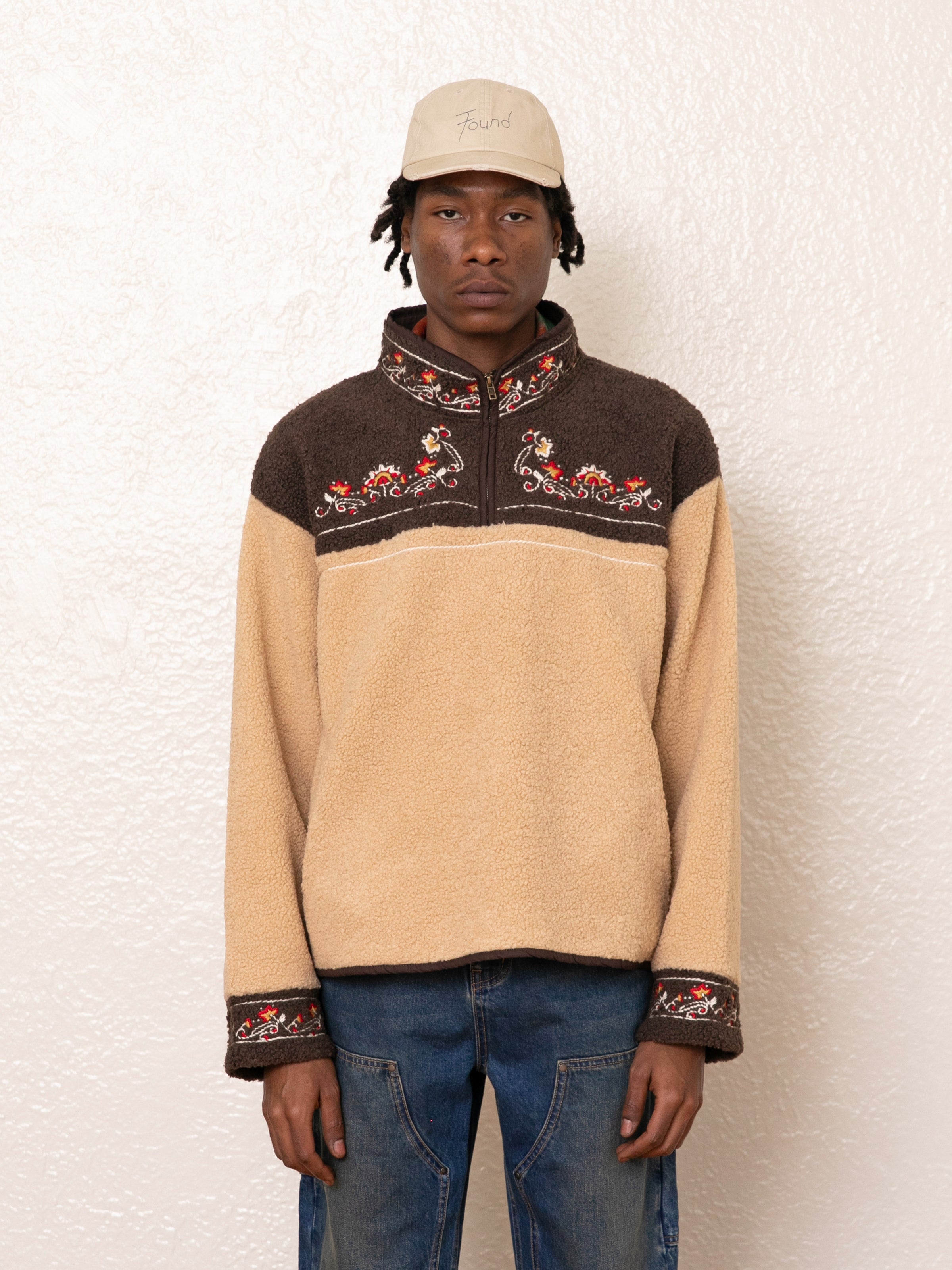 A person in a cap, blue jeans, and the FOUND Pahari Floral Embroidered Sherpa Pullover stands against a textured white wall.