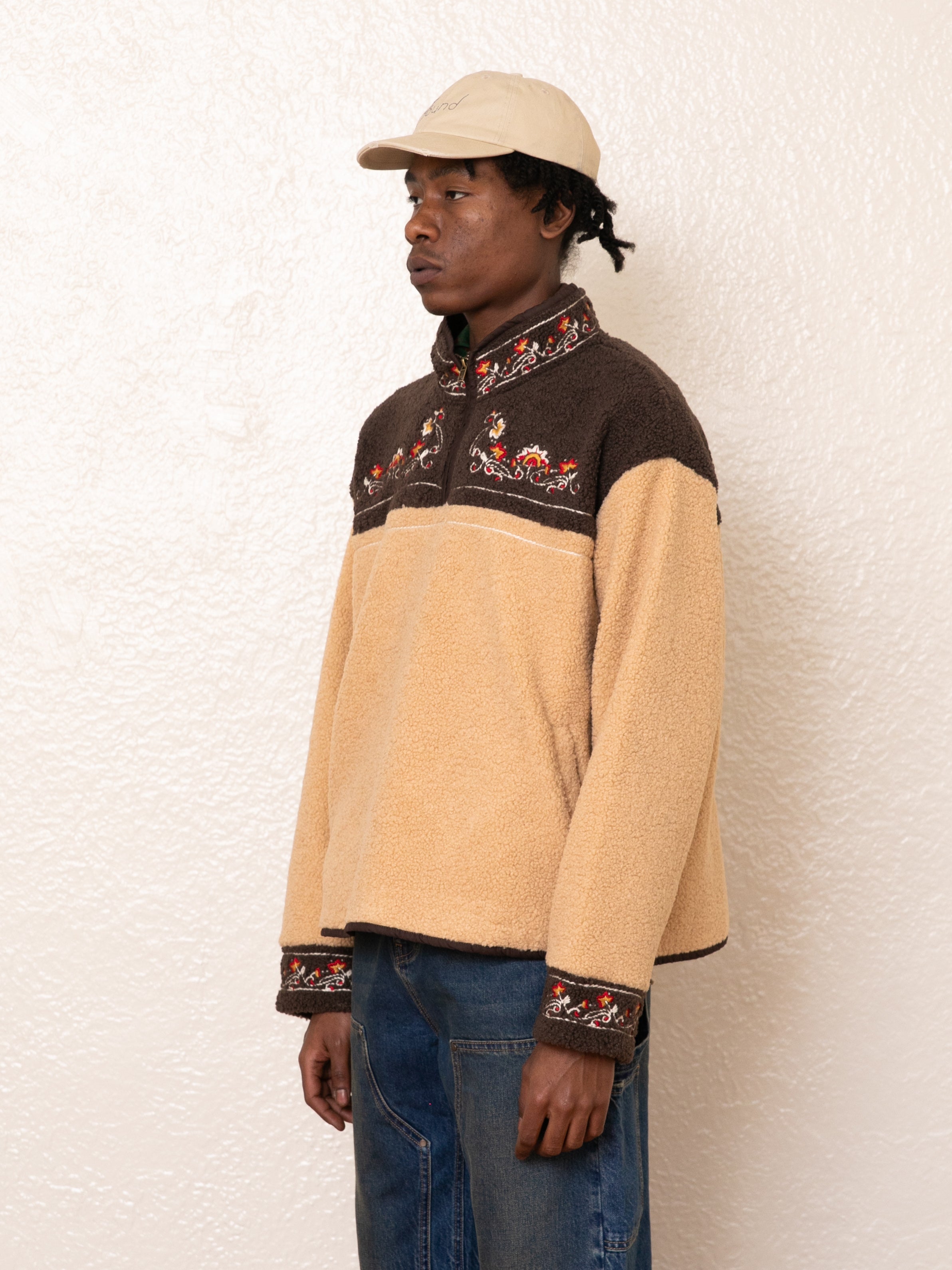 Against a textured white wall, a person wears the FOUND Pahari Floral Embroidered Sherpa Pullover in beige and brown, paired with a beige cap.