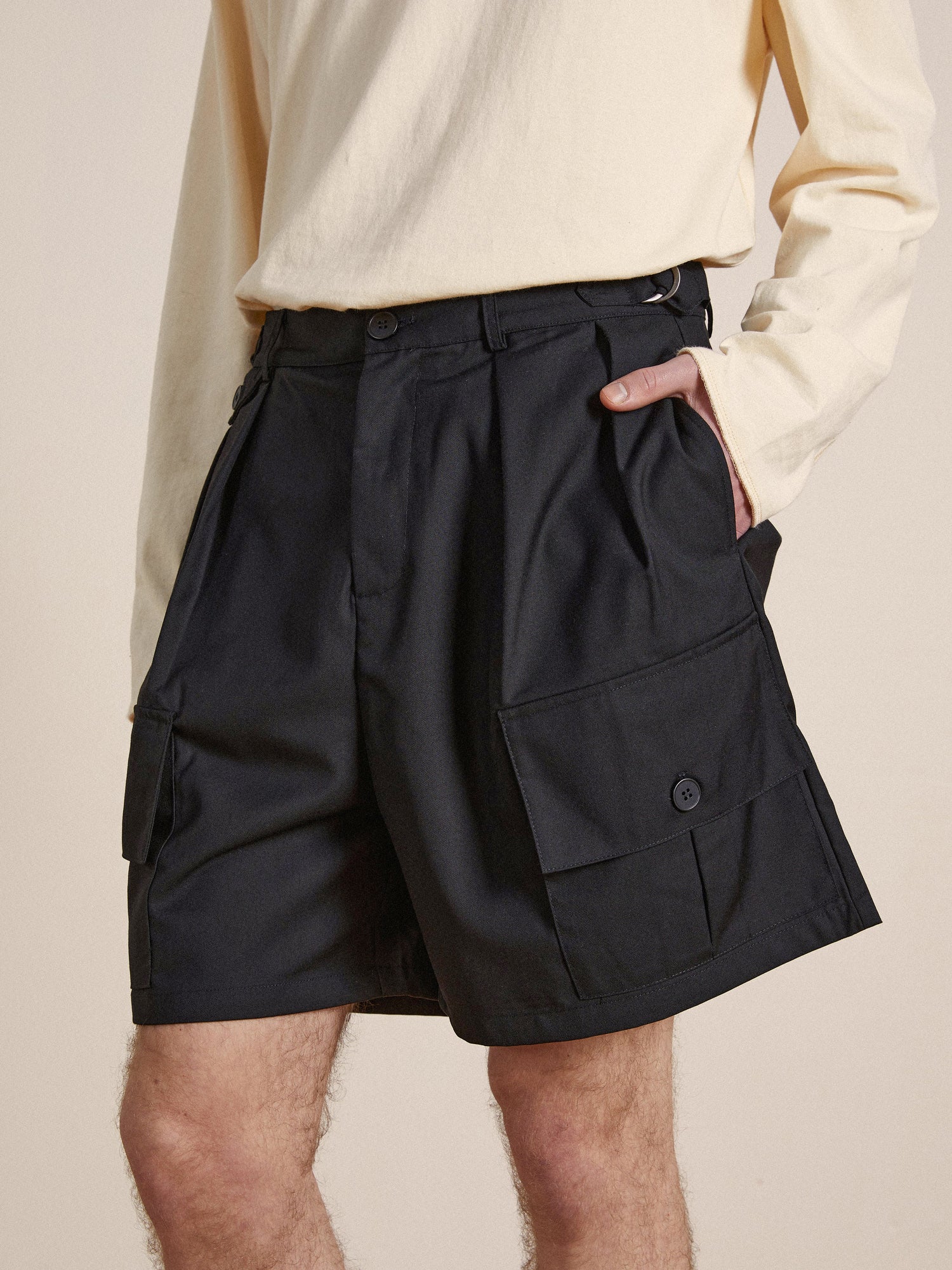 A person wearing a beige top and black Found Pleated Pocket Cargo Shorts stands with one hand in the pocket, focusing on the shorts' details and design.