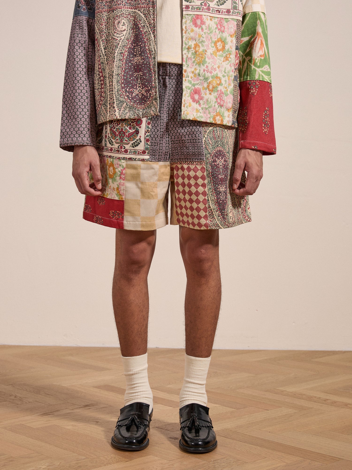 A person wearing a colorful, patchwork-patterned jacket and FOUND's Tapestry Twill Shorts, paired with white socks and black loafers, stands on a wooden floor against a plain background.