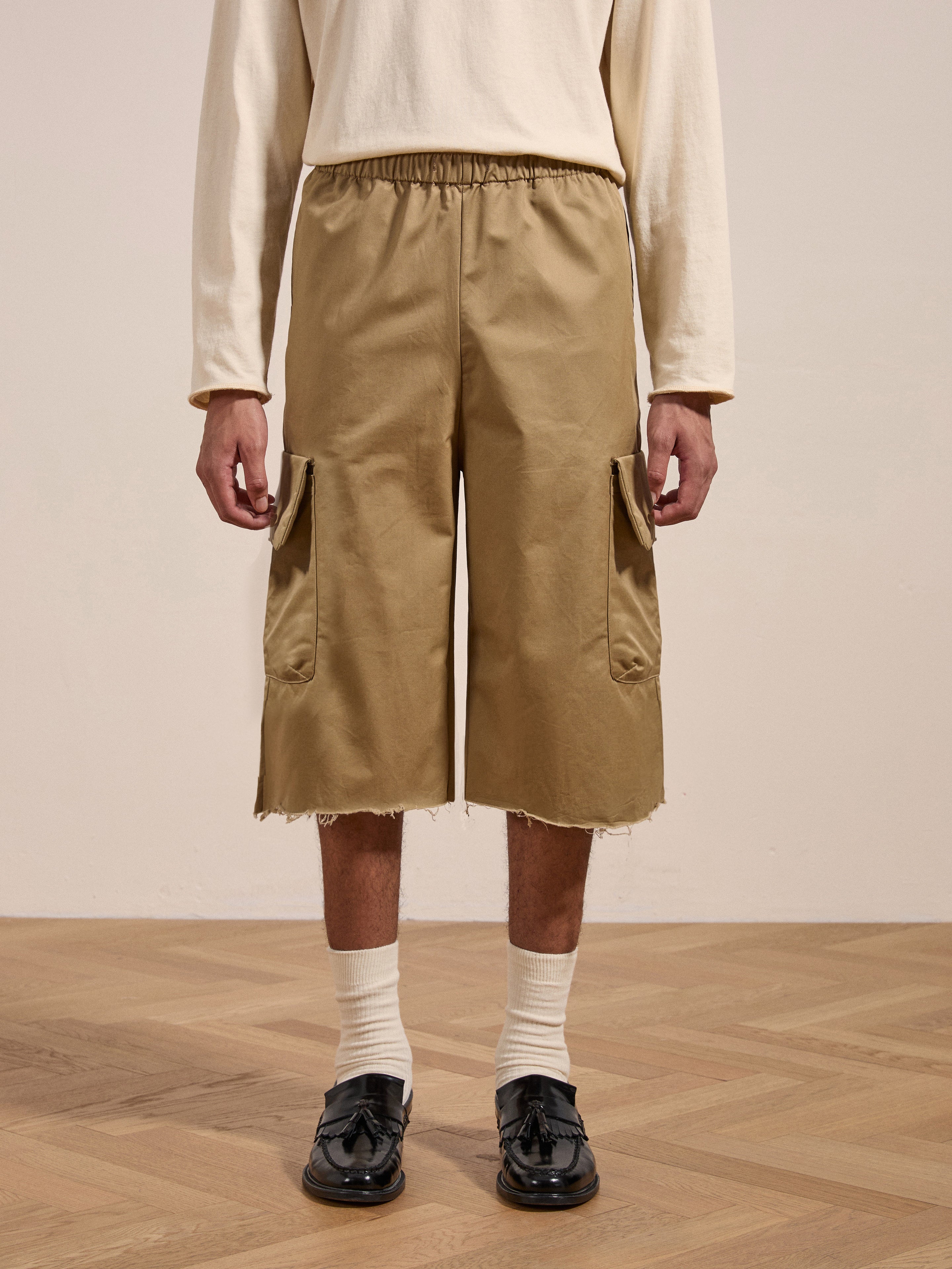 A person is wearing beige Long Twill Cargo Shorts from FOUND with a frayed hem, paired with a beige long-sleeve shirt, white socks, and black loafers. Only the lower half of their body is visible as they stand on a wooden floor.