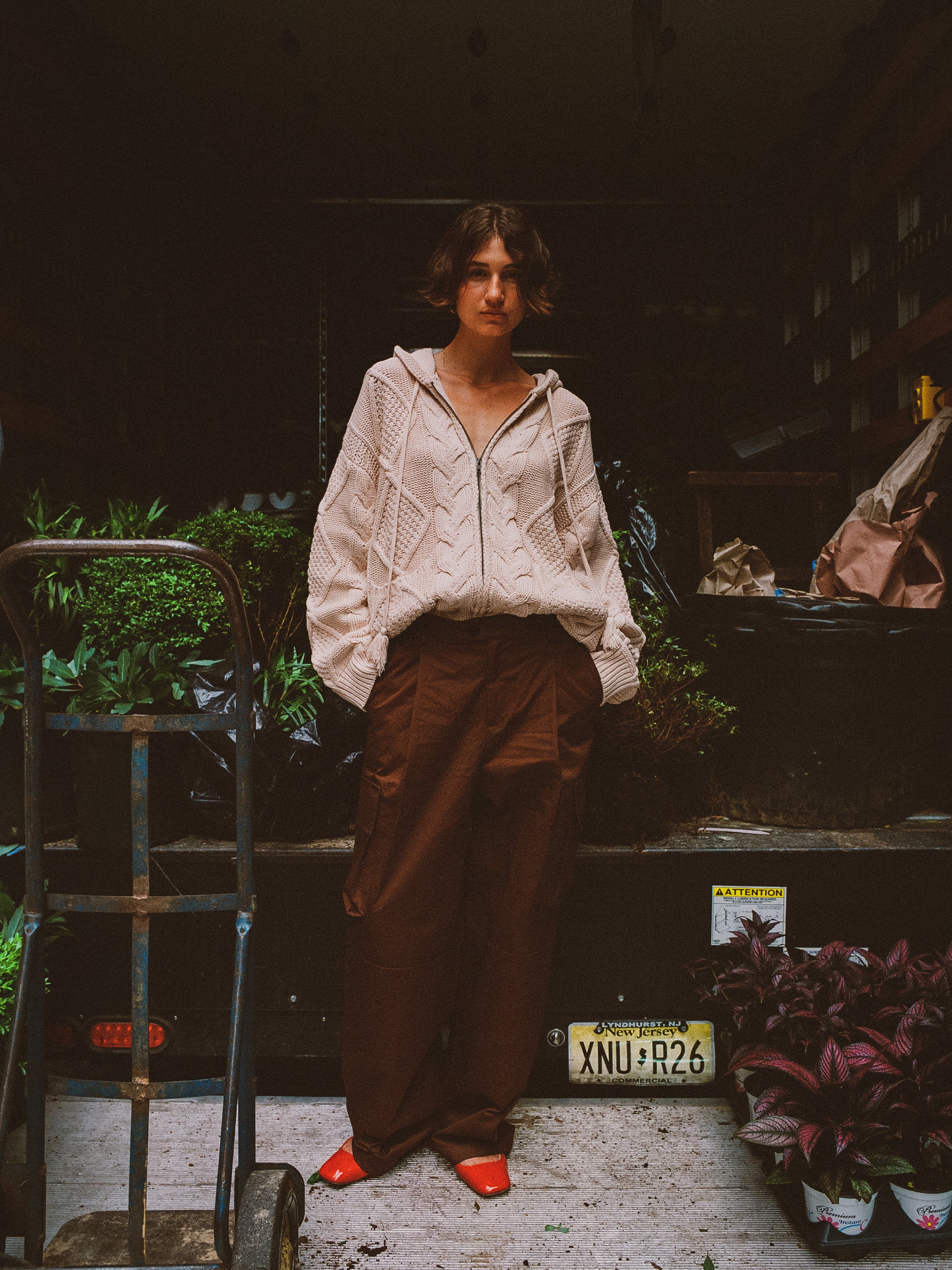 Person standing in a dimly lit space wearing a cozy beige Found Zip Up Cable Knit Hoodie, brown pants, and red shoes, with plants and a dolly cart nearby.