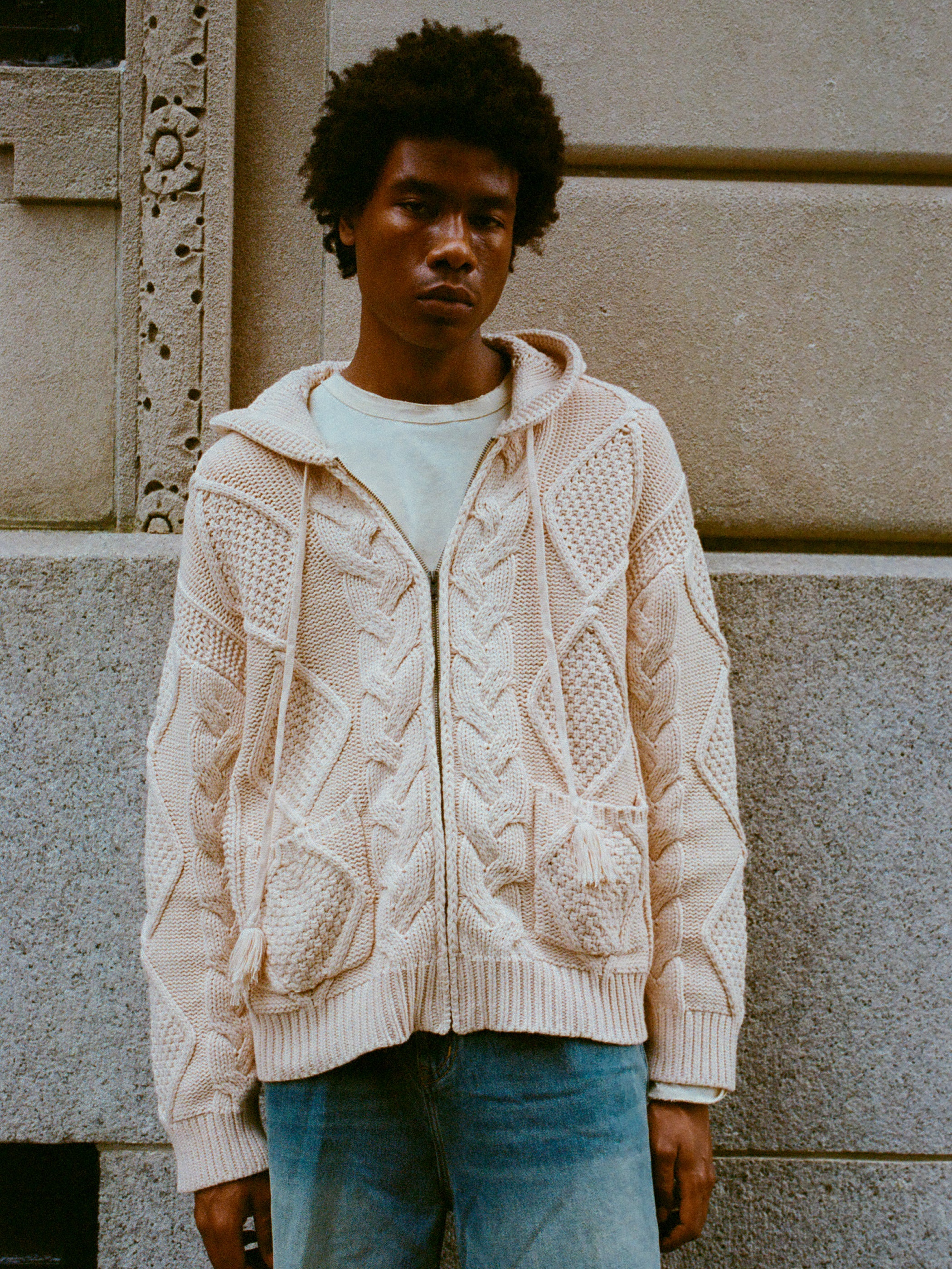 A person with short curly hair stands against a stone wall wearing a cozy Found Zip Up Cable Knit Hoodie and light blue jeans.