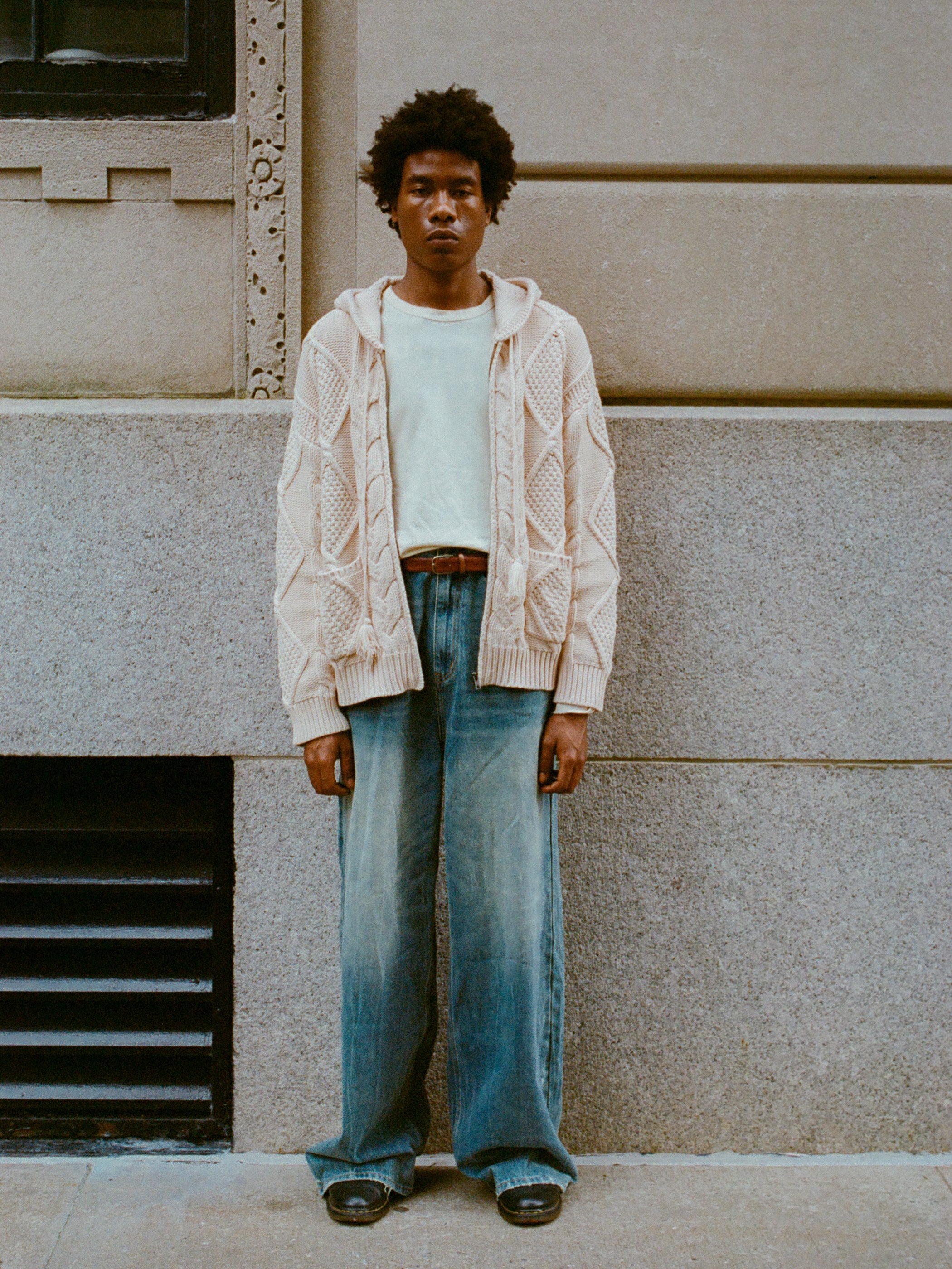 A person with an afro hairstyle is standing against a stone wall, wearing a beige heavyweight cotton blend Zip Up Cable Knit Hoodie by Found over a white T-shirt, blue wide-leg jeans, and dark shoes.