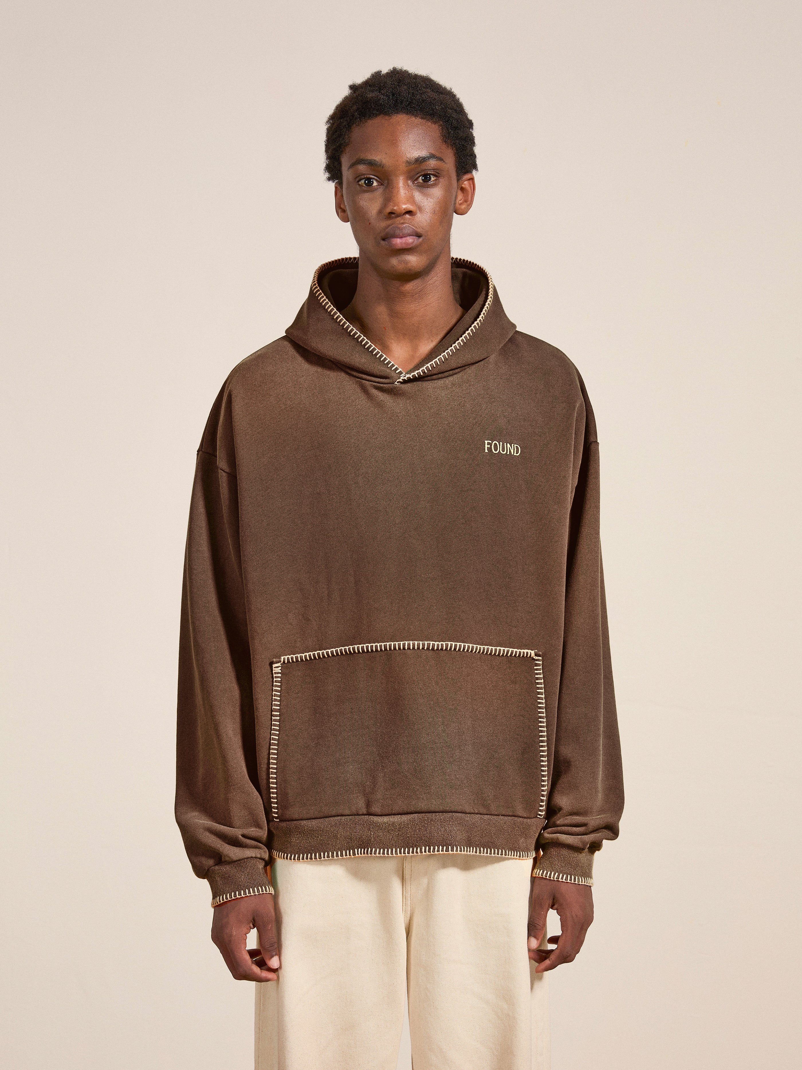 A person stands against a plain background wearing a vintage-style outfit, featuring a brown Stitched Border Distressed Logo Hoodie by FOUND, paired with light-colored pants.