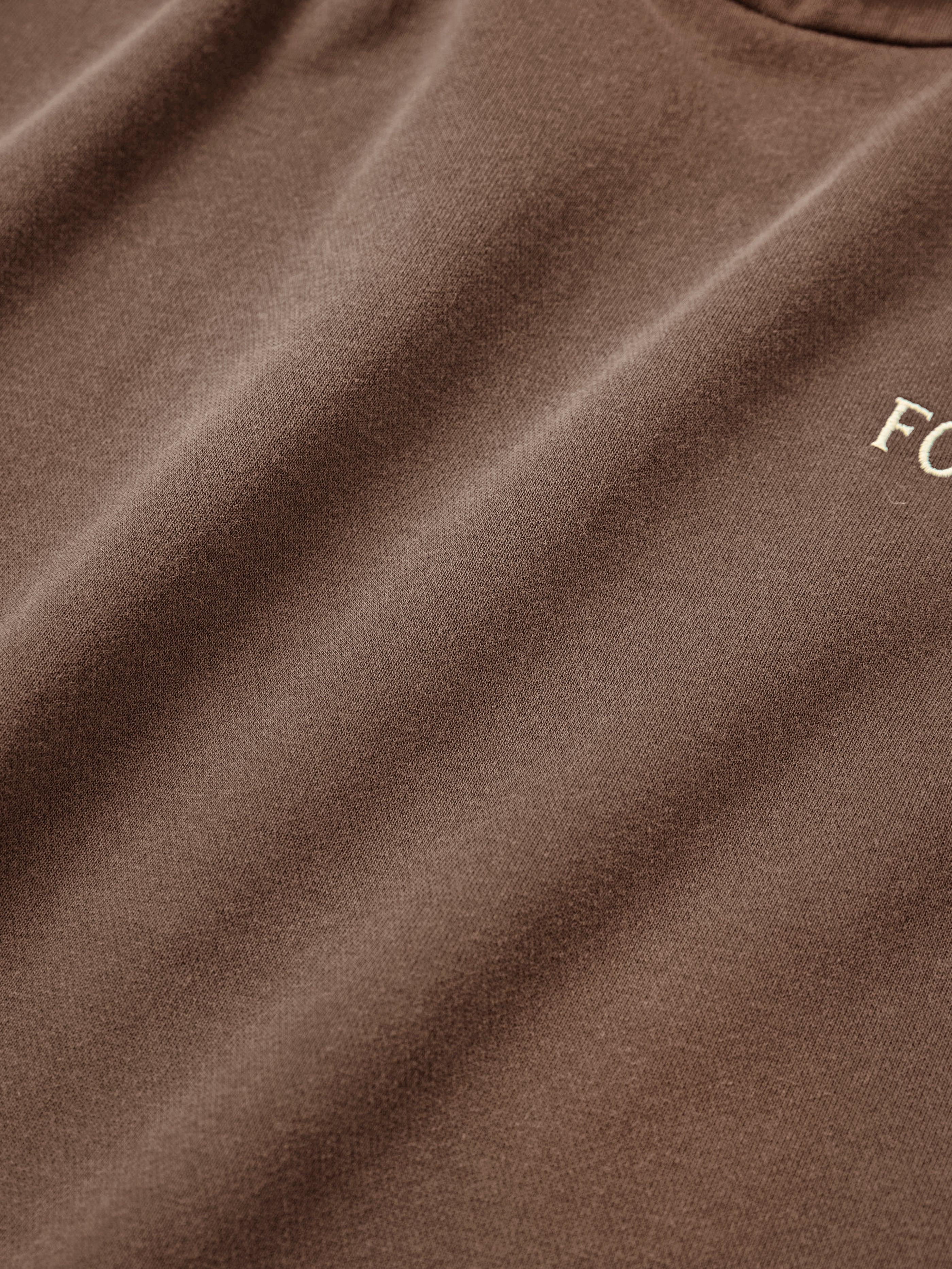 A close-up of a brown fabric with subtle folds and the partial word "FO" embroidered in white evokes a vintage feel, reminiscent of the oversized Stitched Border Distressed Logo Hoodie crafted by FOUND.