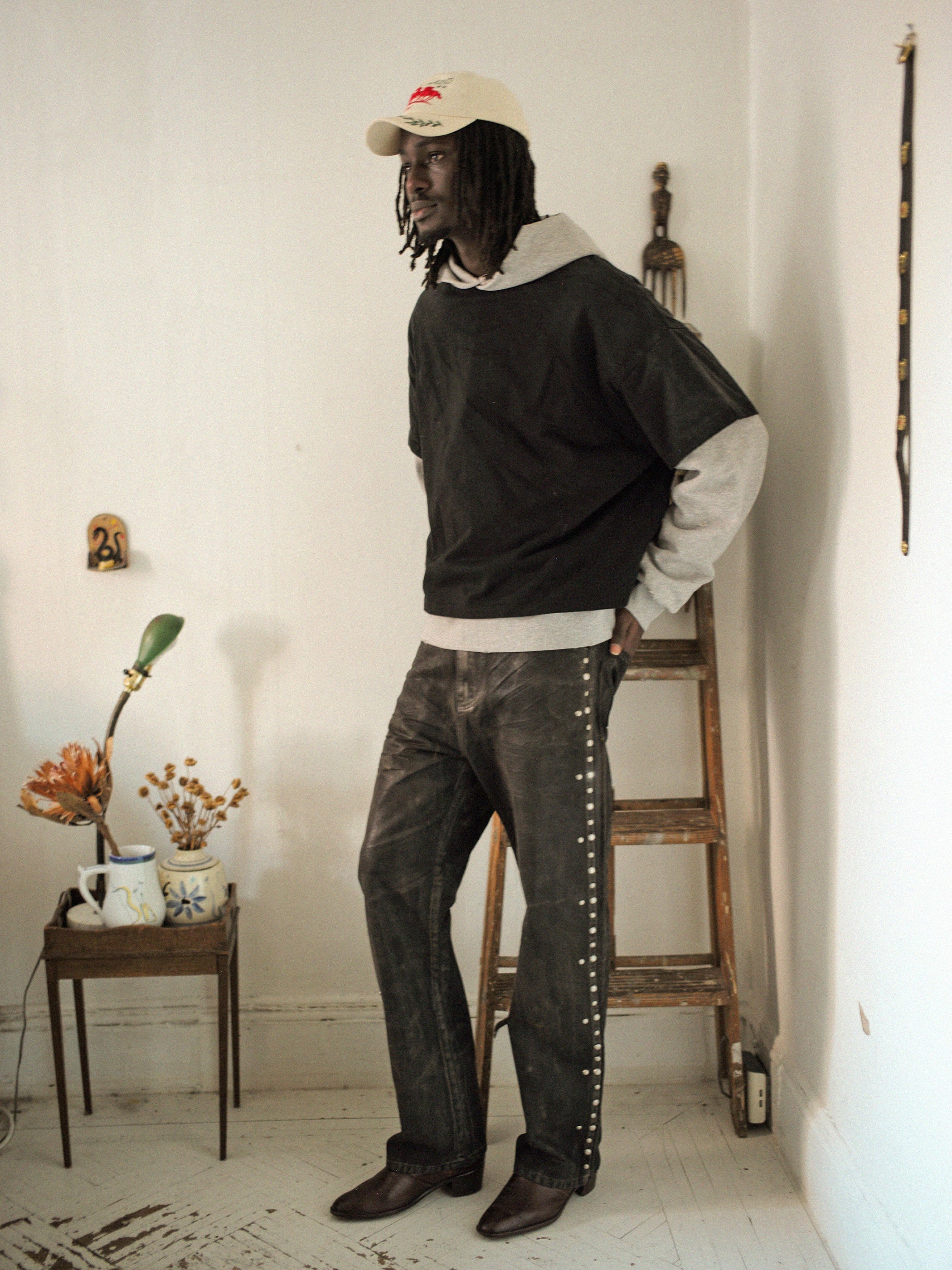 In a room with minimalistic decor, a person in a vintage look stands beside a wooden ladder, wearing the Found Double Layer Hoodie next to a small table with flowers and a cup on their left.