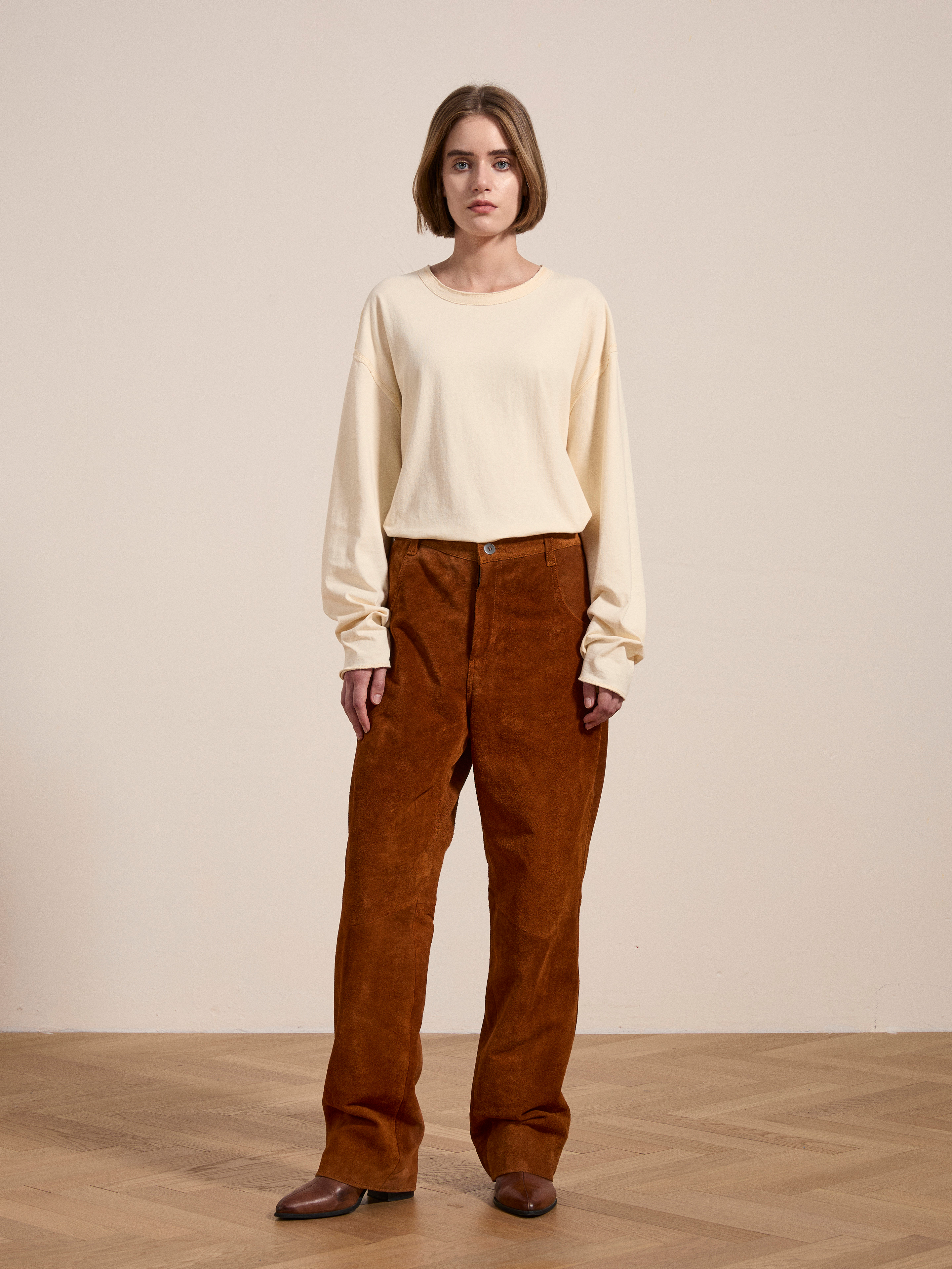 A person stands on a wooden floor, wearing a cream long-sleeve shirt, ethically sourced brown suede moto pants by FOUND, and brown shoes, set against a plain beige background.