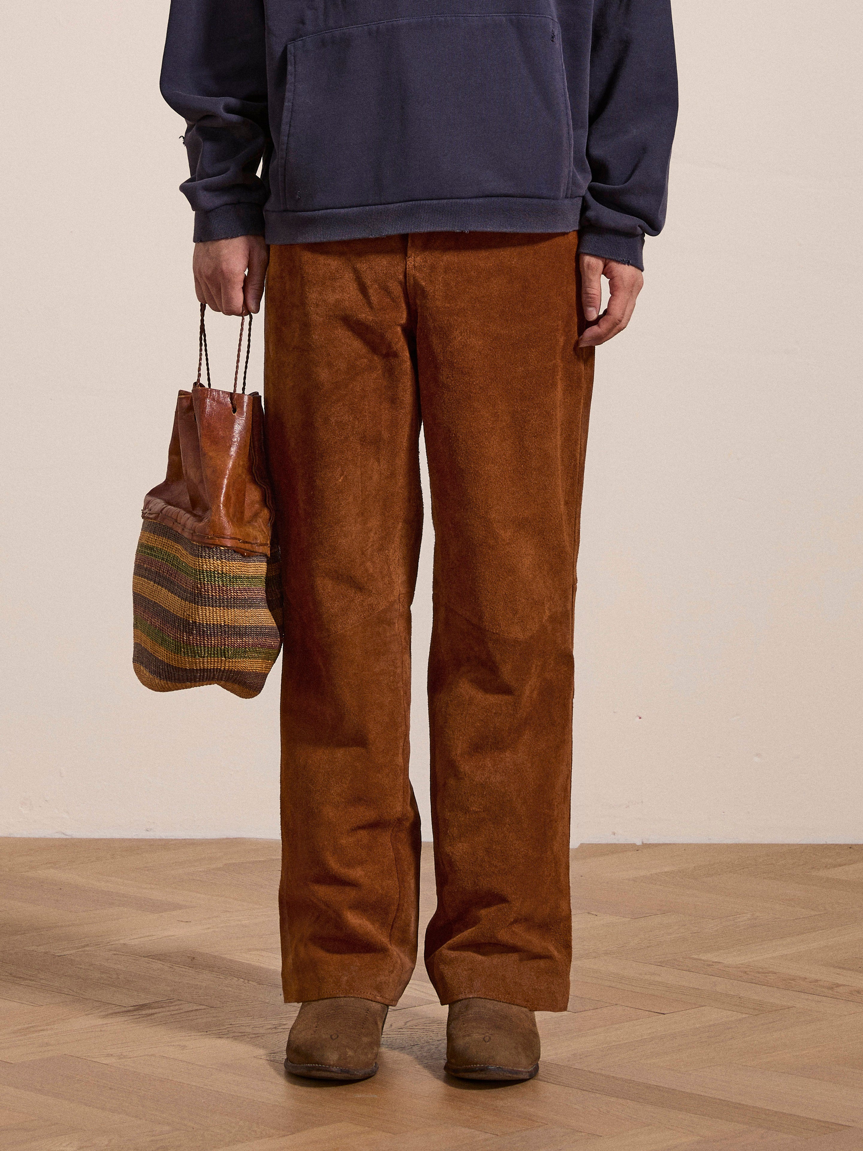 A person standing on a wooden floor, dressed in FOUND's Suede Moto Pants, a dark blue sweatshirt, brown shoes, and holding a striped bag.