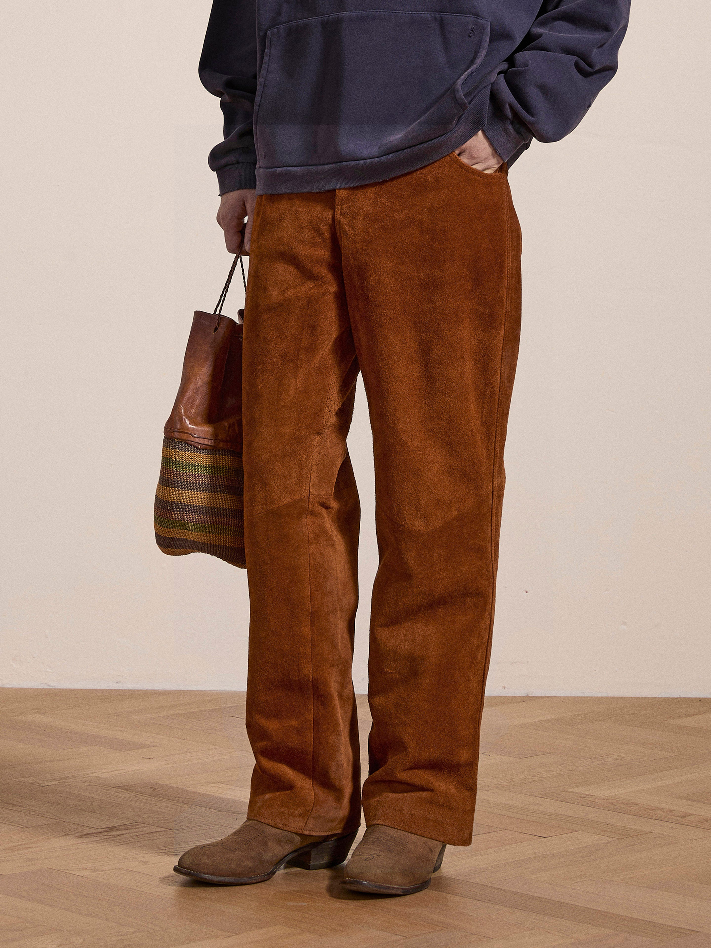 A person stands wearing Suede Moto Pants from FOUND in a rust hue, paired with a navy sweater and brown ethically sourced boots, holding a brown and green striped bag.