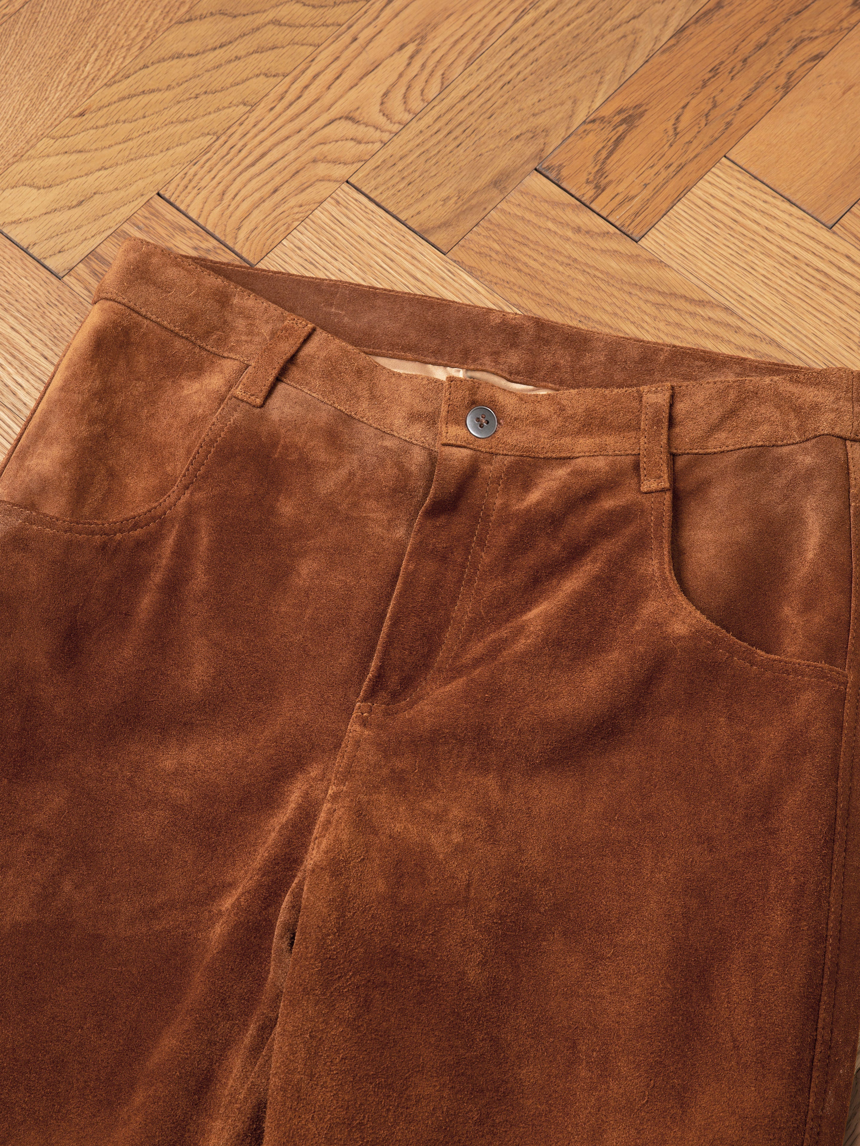 Close-up of FOUND's Suede Moto Pants, crafted from heavyweight nappy suede and ethically sourced materials, featuring a single button and zip closure, displayed on a wooden herringbone floor.