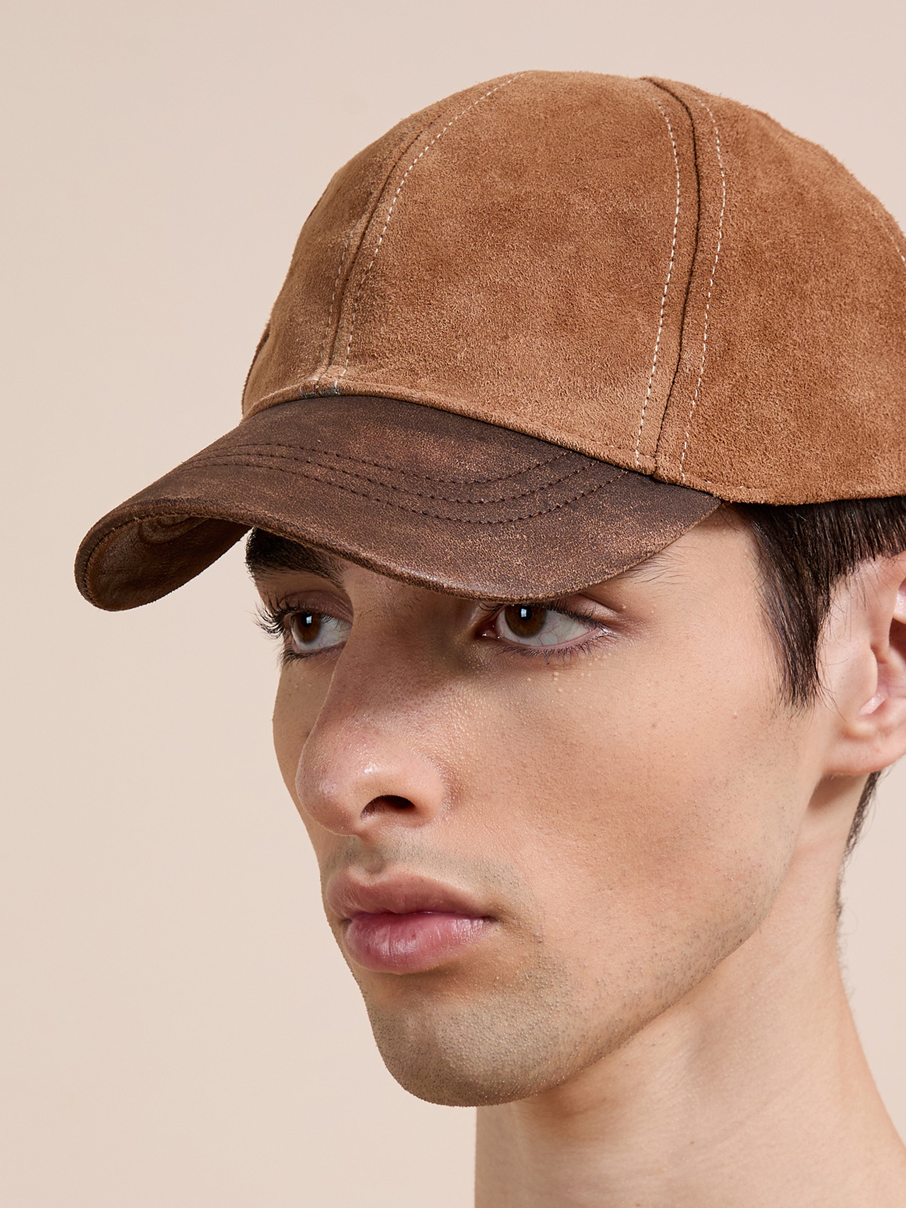A young person models the Found Suede Leather Contrast Distressed Cap, featuring brown suede and an adjustable leather strap, set against a plain beige background.