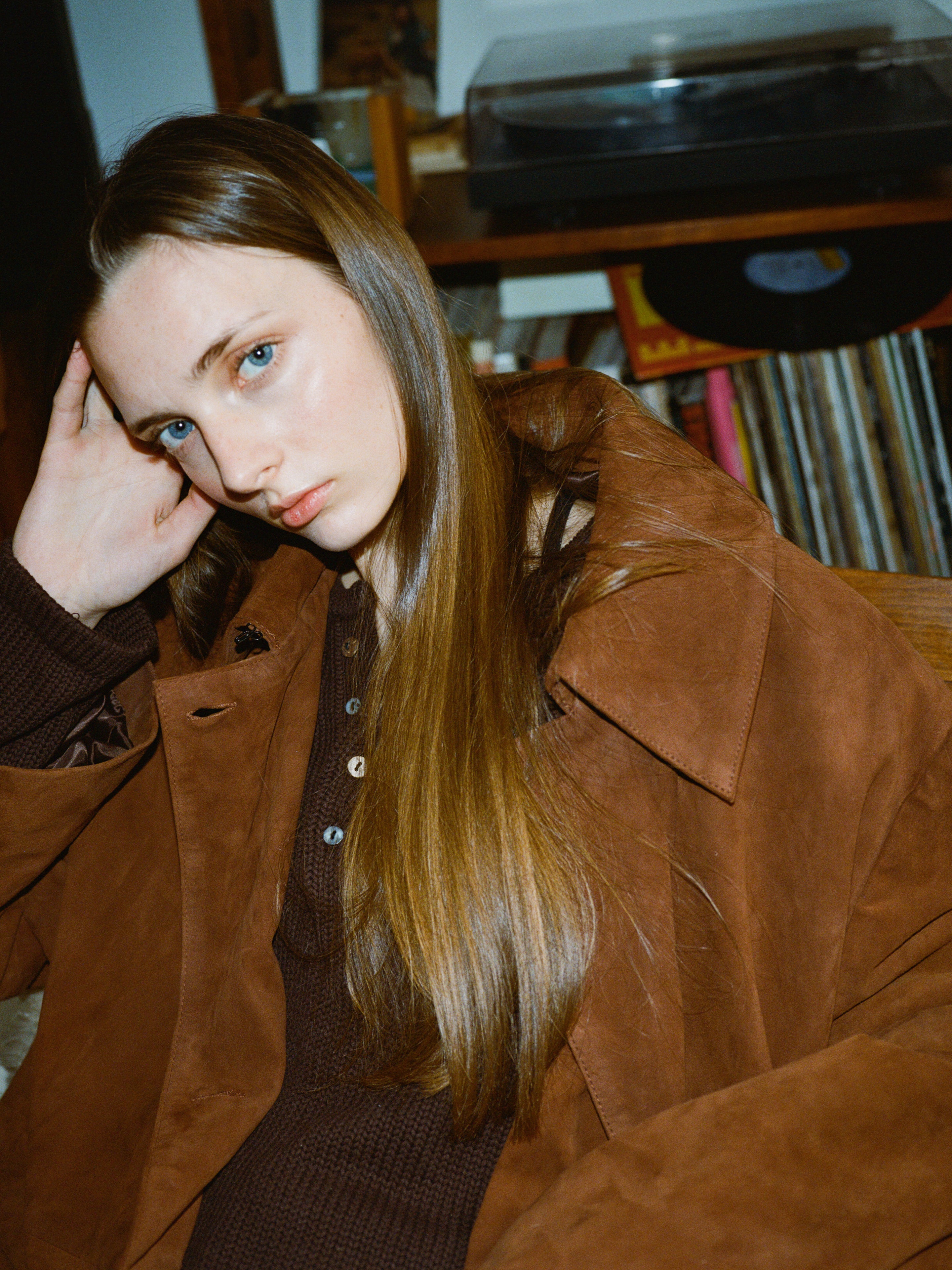 In a cozy indoor setting, someone with long hair wearing a FOUND Double Breasted Suede Leather Jacket sits near a record shelf and turntable, gazing at the camera.