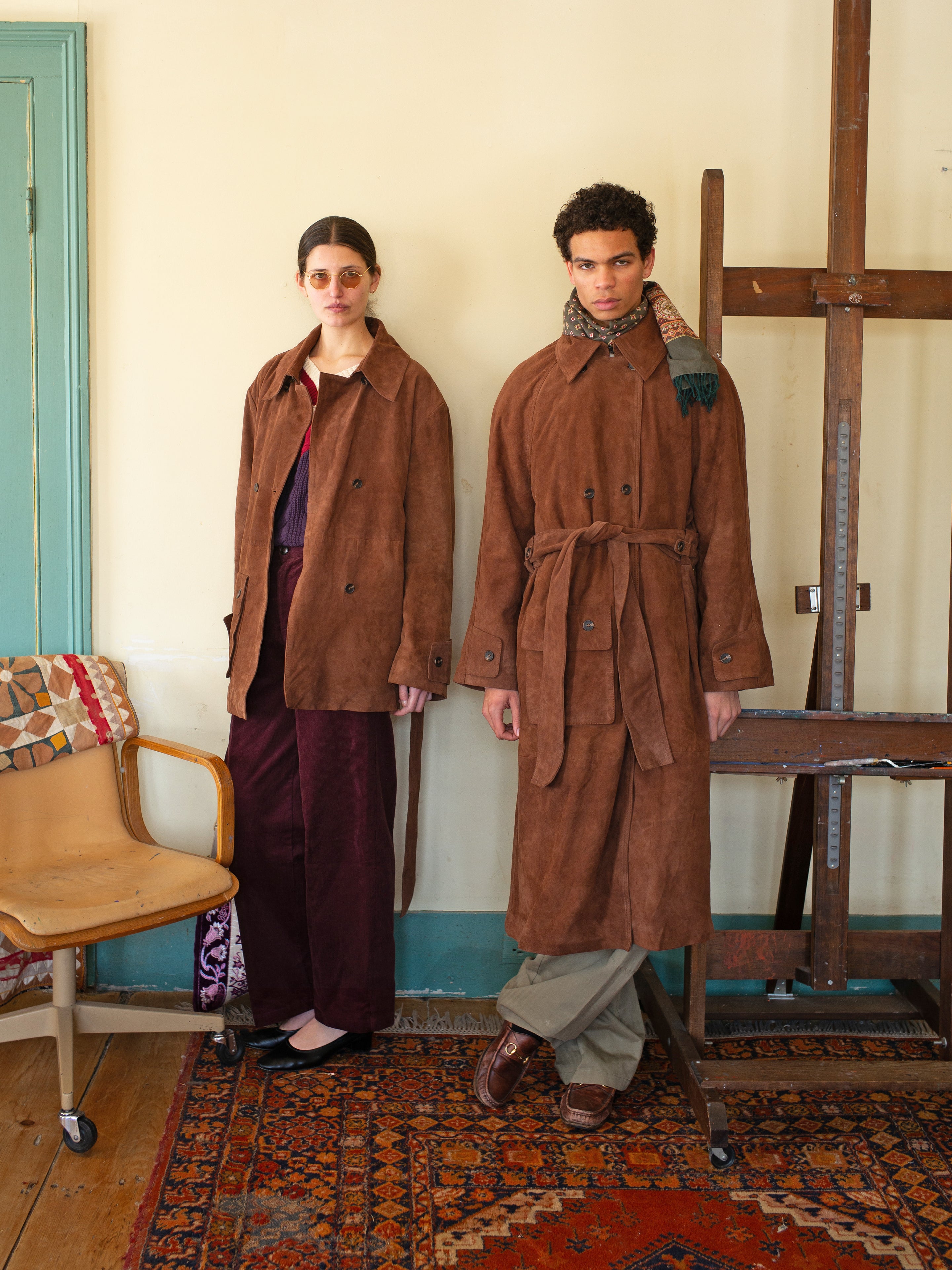 Two individuals in brown coats, one wearing FOUND's Suede Leather Trench Coat and the other with a scarf, stand in a room adorned with a wooden chair, large easel, and an elegantly patterned rug—showcasing ethically crafted unisex fashion from Pakistan.