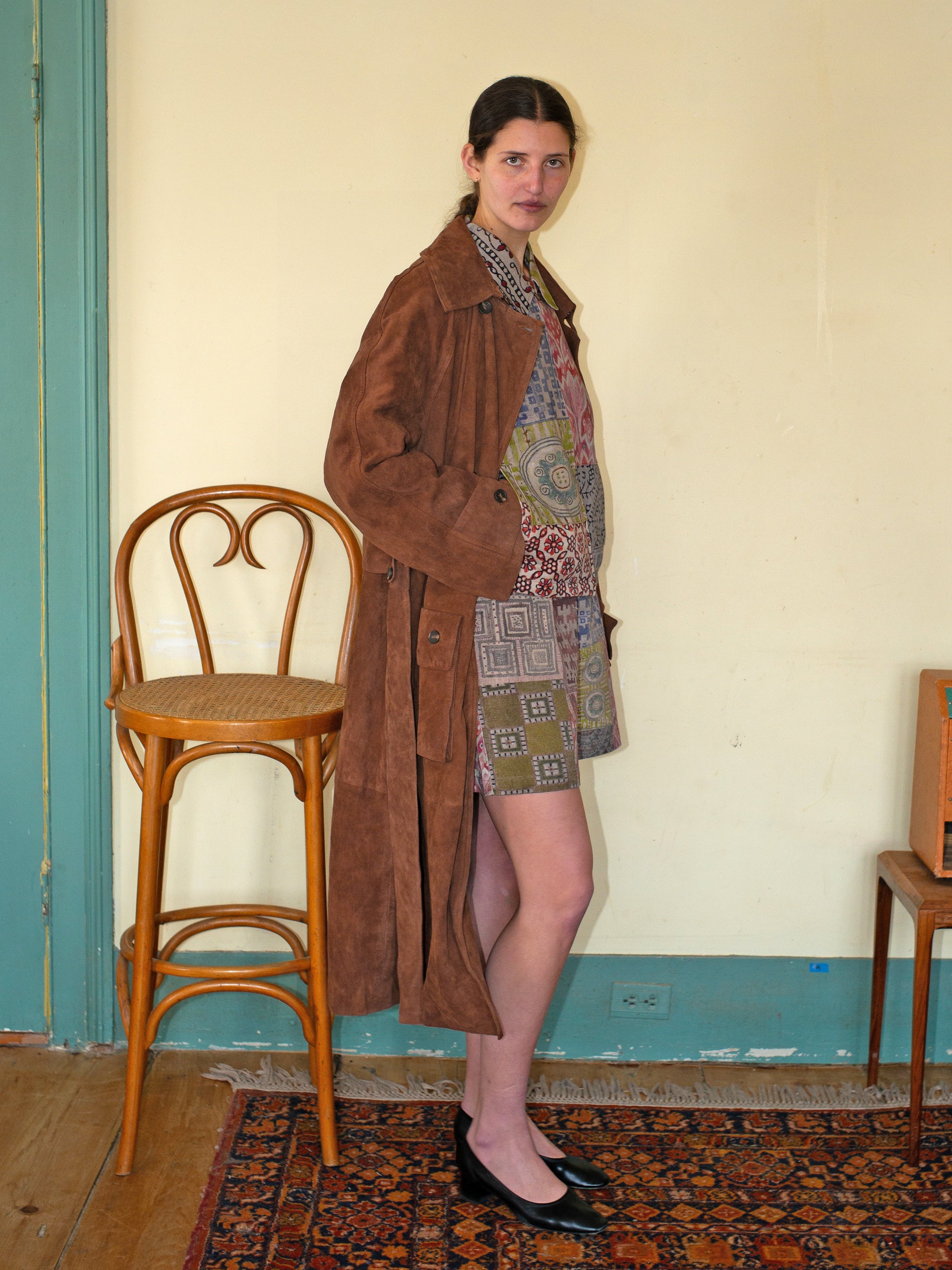 Indoors, a person in a FOUND Suede Leather Trench Coat, ethically crafted in Pakistan, pairs the outfit with a patterned dress and black shoes. They stand beside a wooden chair on top of a rug.