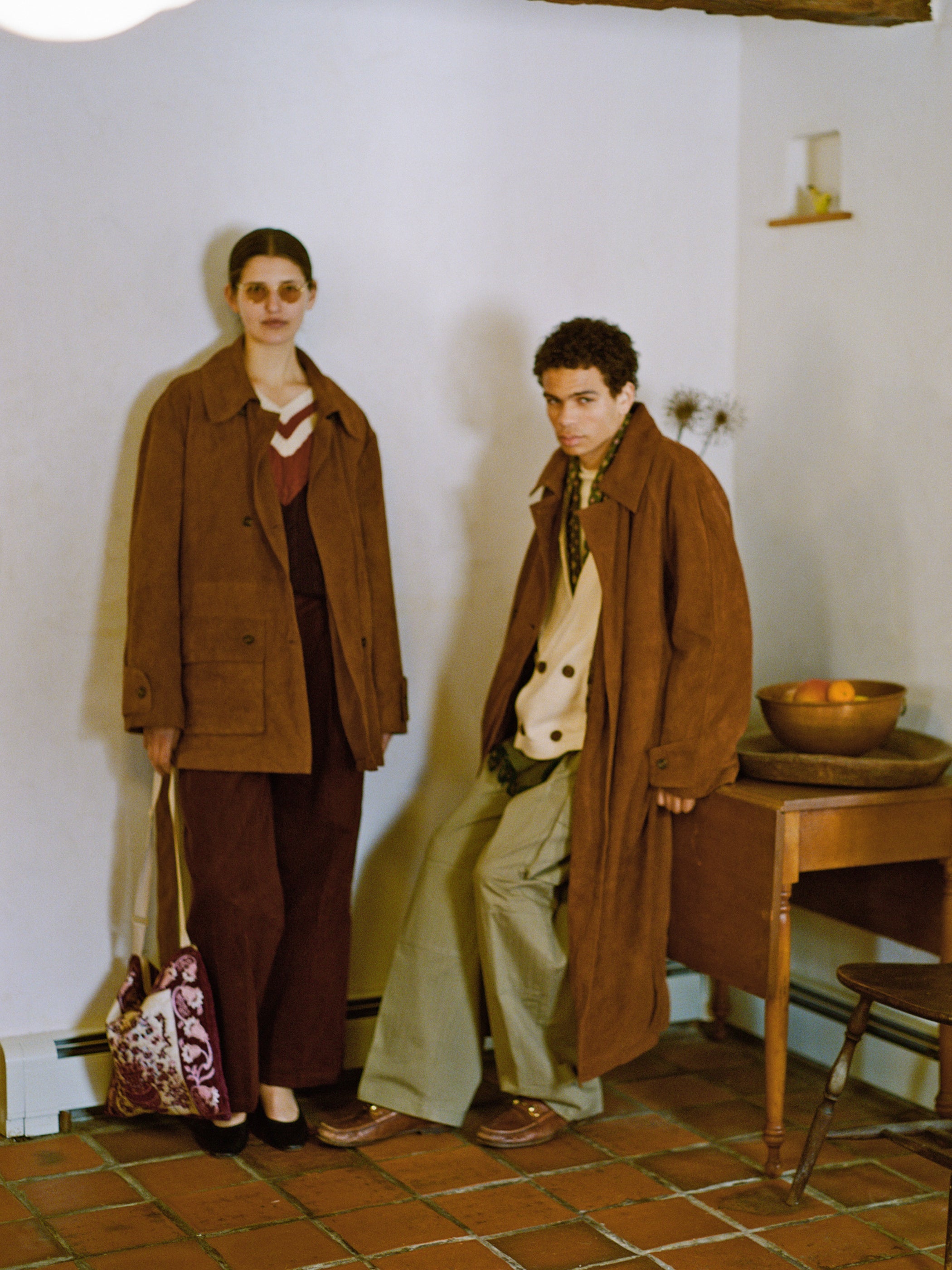 Two people in FOUND unisex Suede Leather Trench Coats and sunglasses stand by a small wooden table with fruit. Ethically crafted in Pakistan, these coats add sophistication to any setting.