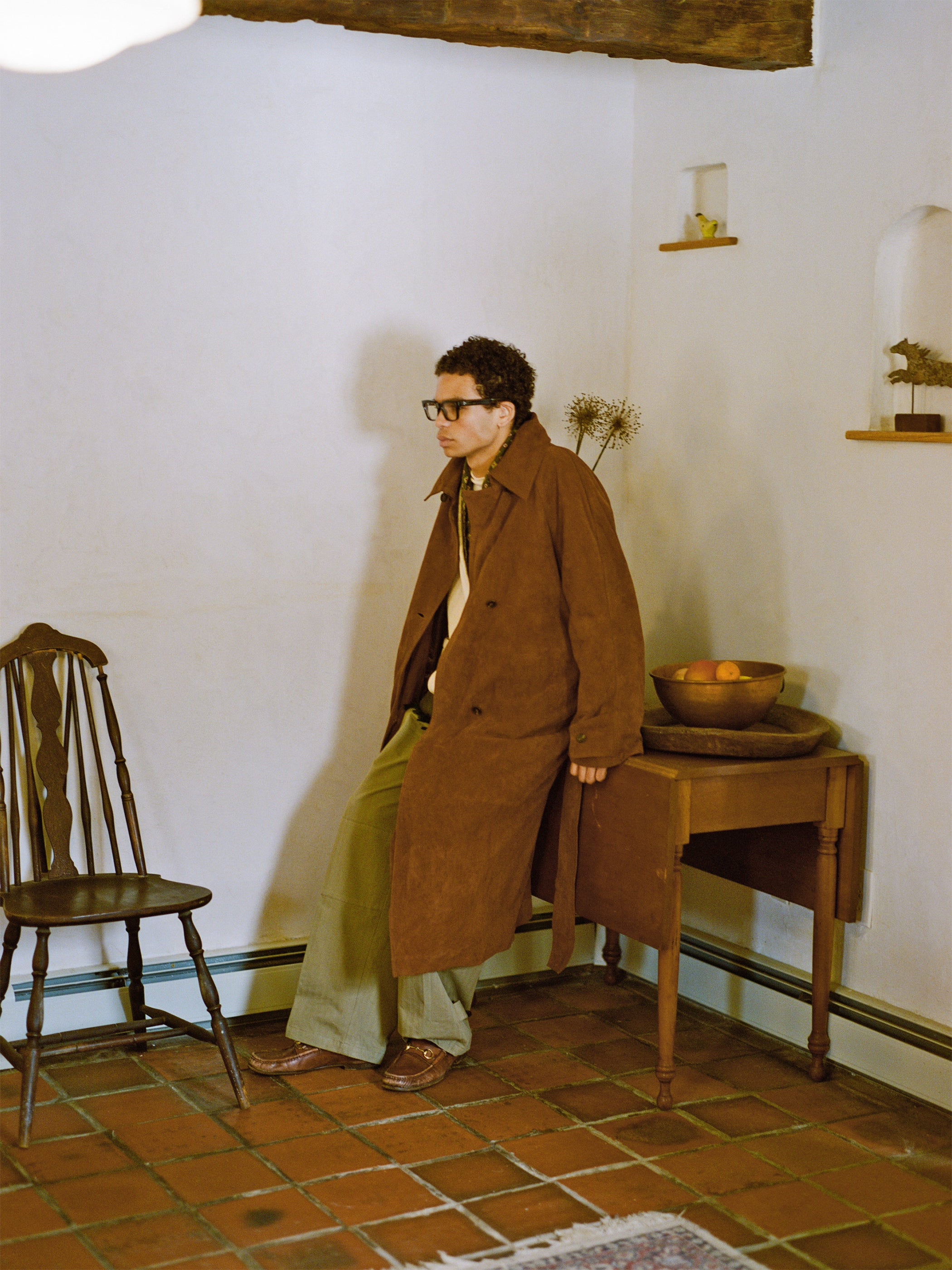 A person wearing FOUND's ethically crafted Suede Leather Trench Coat and glasses leans against a small table with a bowl of oranges in a rustic room, highlighting the tiled floor and wooden chair.