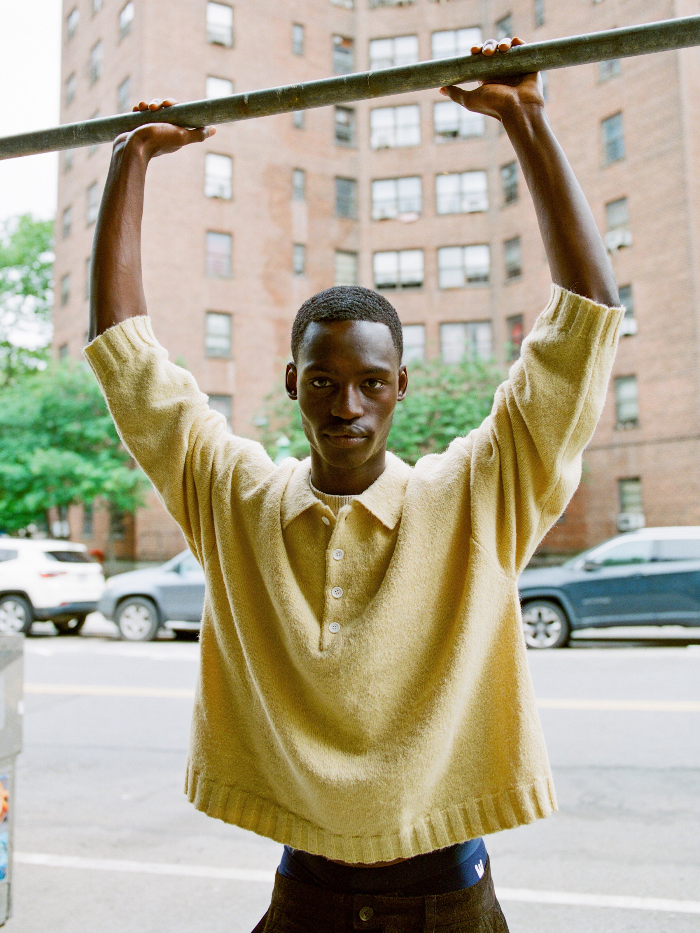 A person wears the FOUND Knit LS Polo Sweater in yellow, shown outdoors holding a metal bar, with brick apartments and parked cars behind them. The sweater is made from soft, brushed knit fabric for comfort and style.