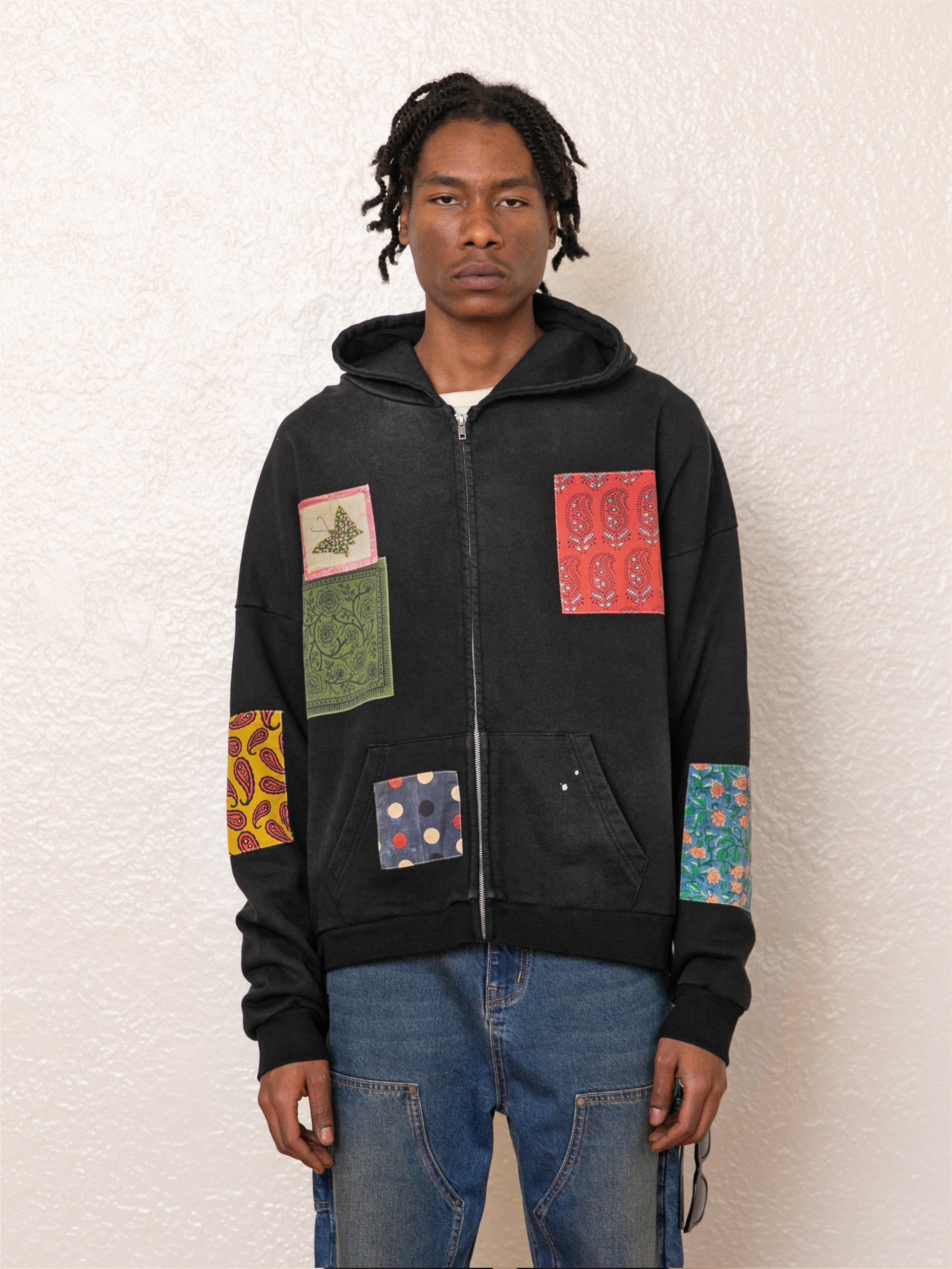 A person in a FOUND Tapestry Patchwork Zip Up Hoodie, featuring a vintage-inspired oversized fit with colorful designs, stands against a textured white wall.