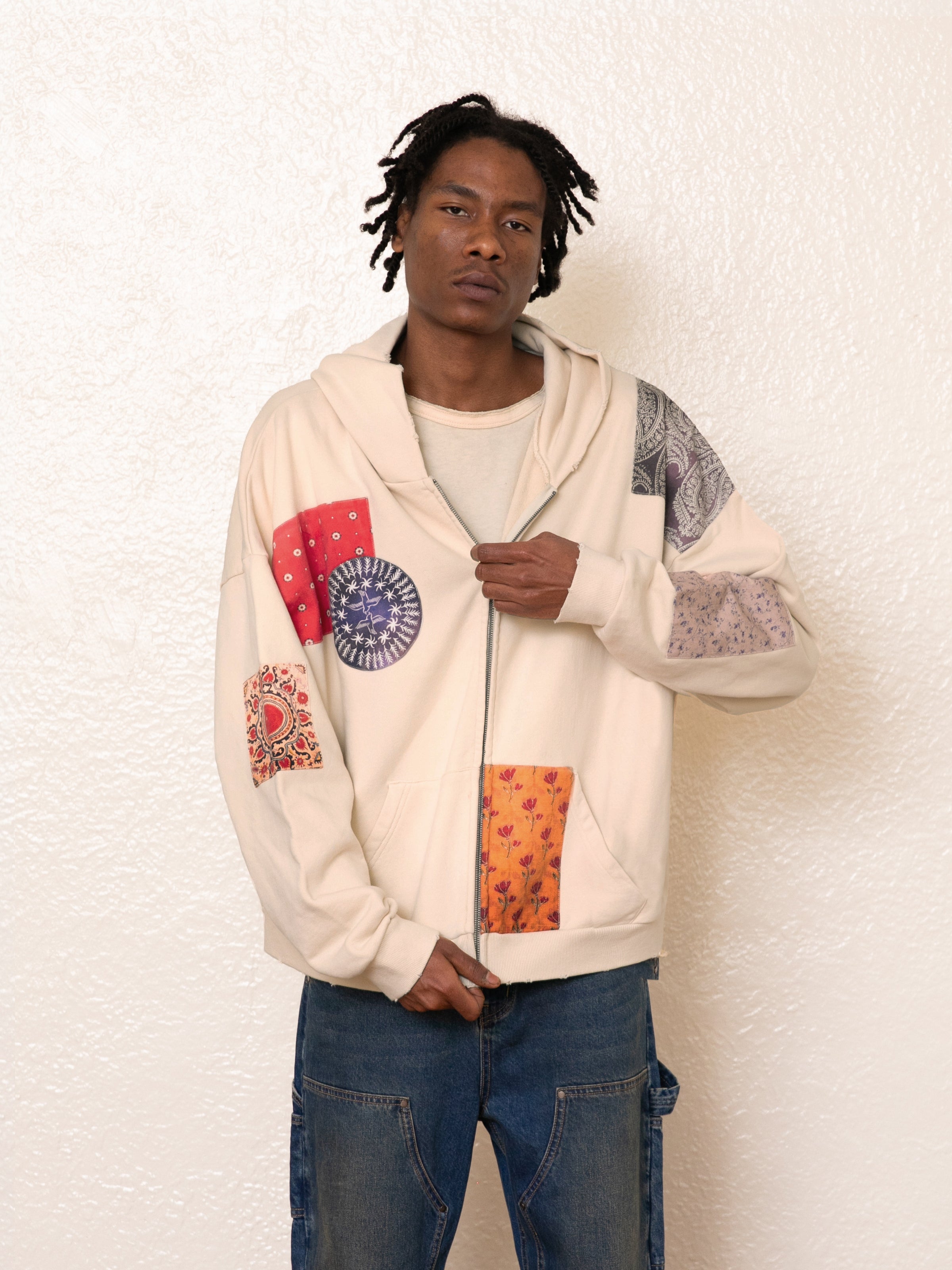 A person wearing a FOUND Tapestry Patchwork Zip Up Hoodie and jeans stands against a textured white wall.