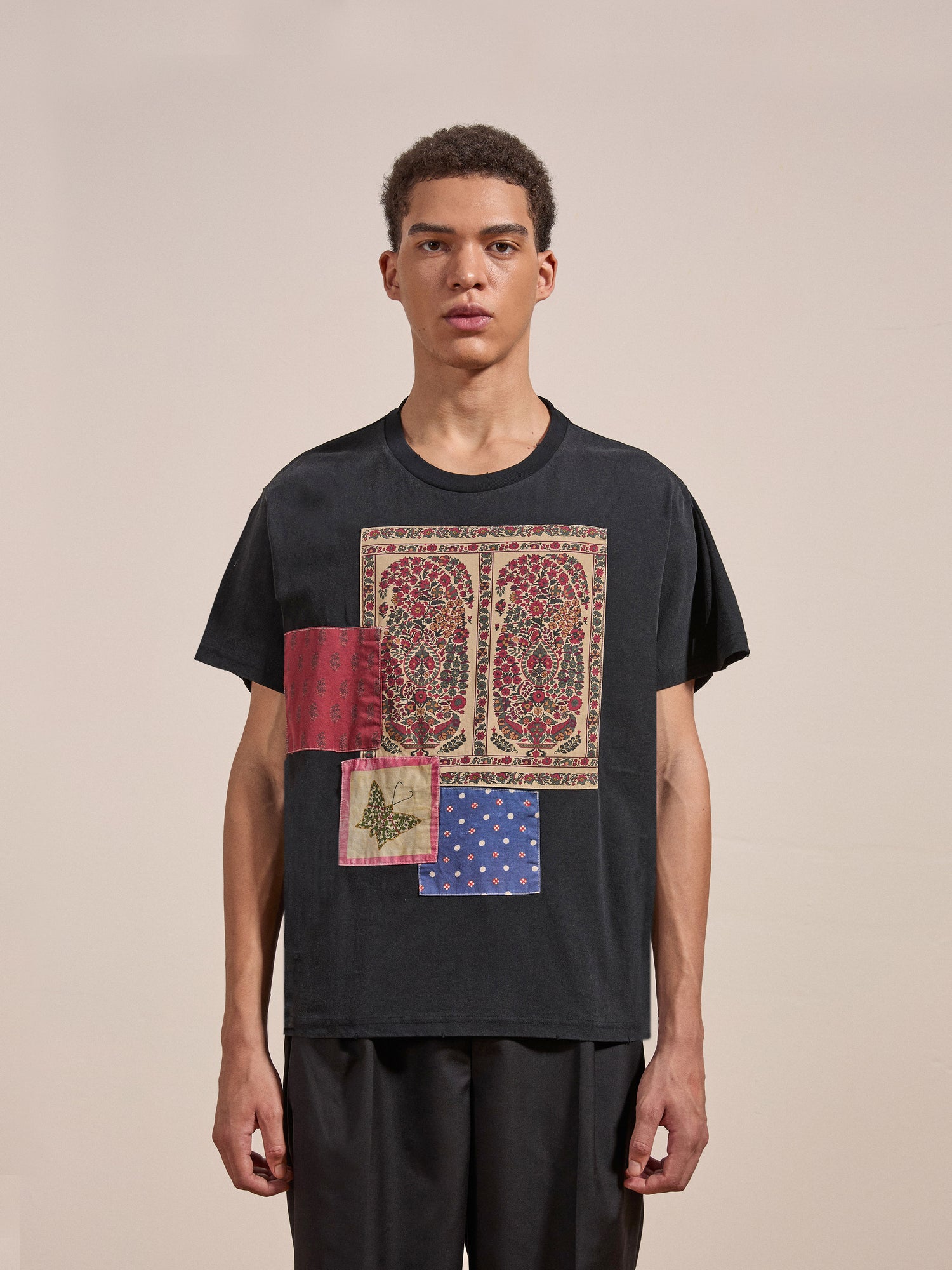 A person stands against a plain background wearing the Patchwork Tee from FOUND, featuring colorful, patterned patches, and black pants.