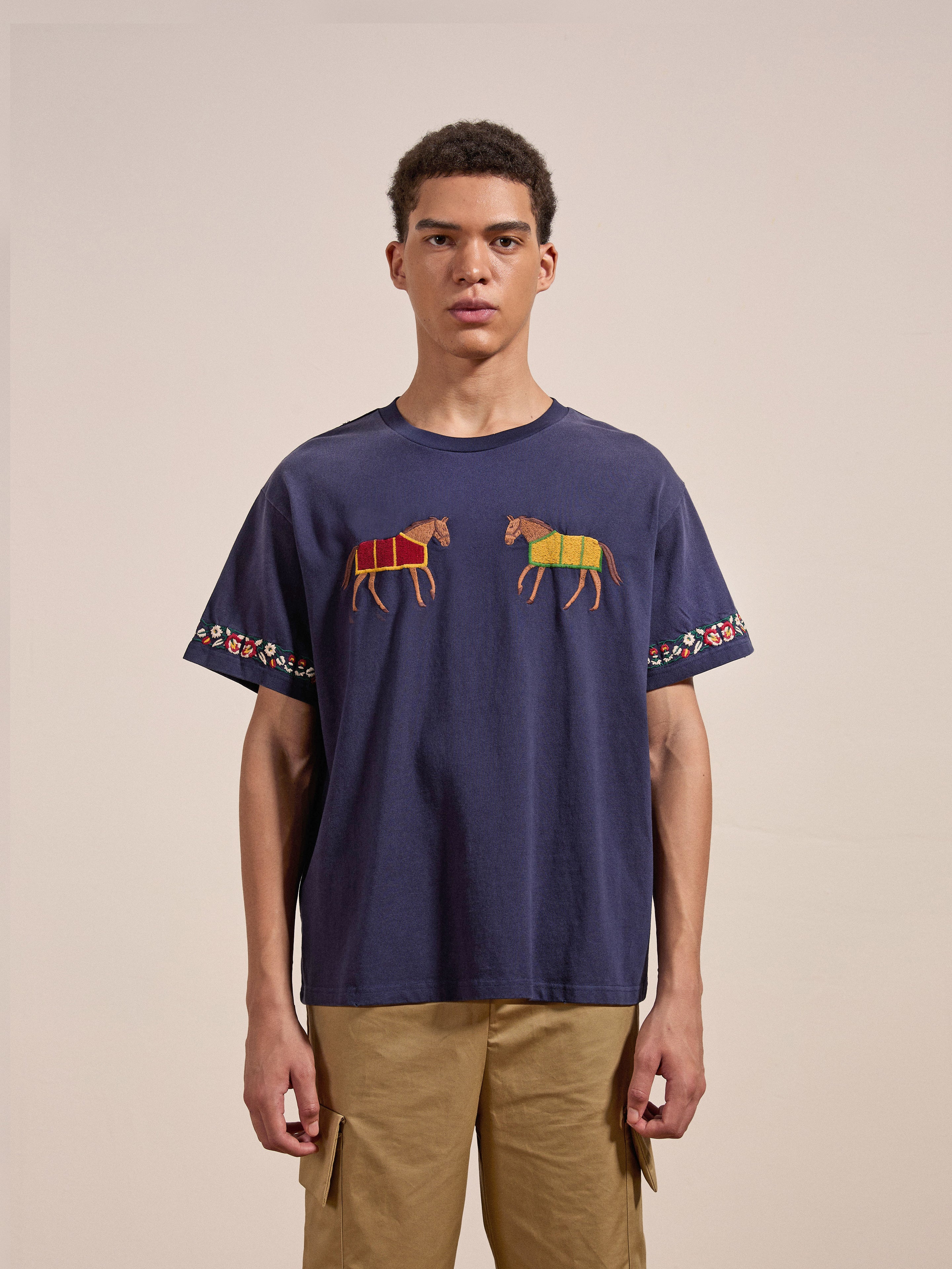 A young person is wearing the FOUND Horse Equine Tee, a blue t-shirt featuring two embroidered horse designs and decorative patterns on the sleeves. They are also dressed in beige pants, with a plain and neutral background complementing their outfit.