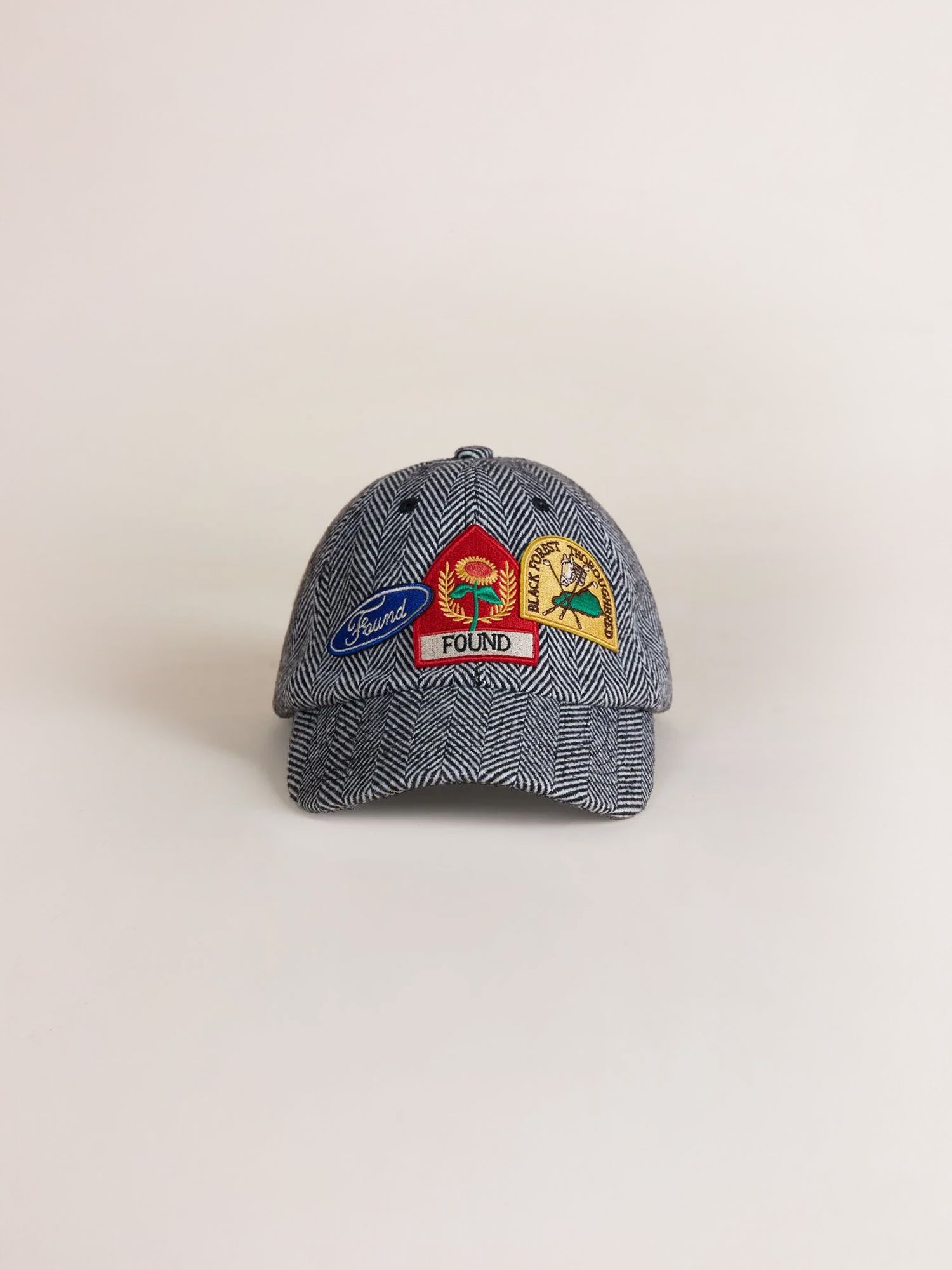 The Herringbone Tweed Patch Cap by Found is a gray cap adorned with vintage patches on the front, showcasing various designs including a bird, a sun, and a tree along with the word 