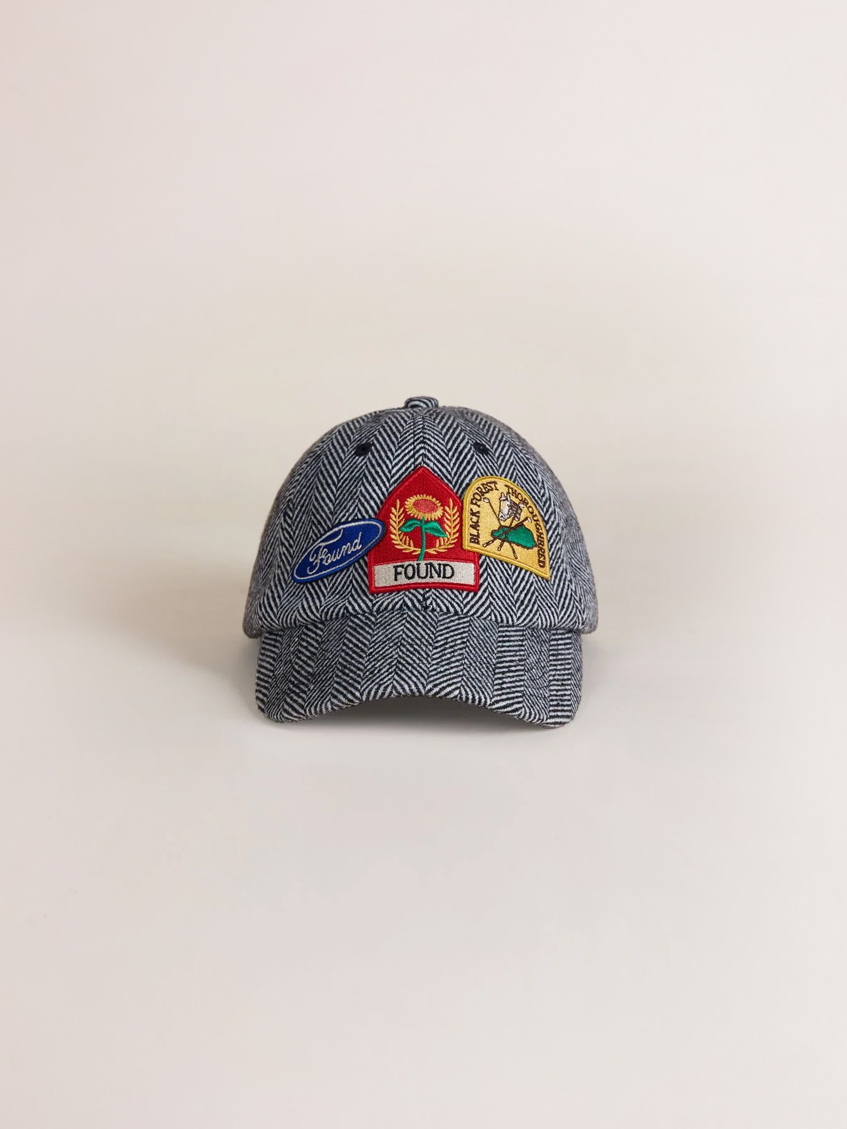 The Herringbone Tweed Patch Cap by Found is a gray cap adorned with vintage patches on the front, showcasing various designs including a bird, a sun, and a tree along with the word "Found".