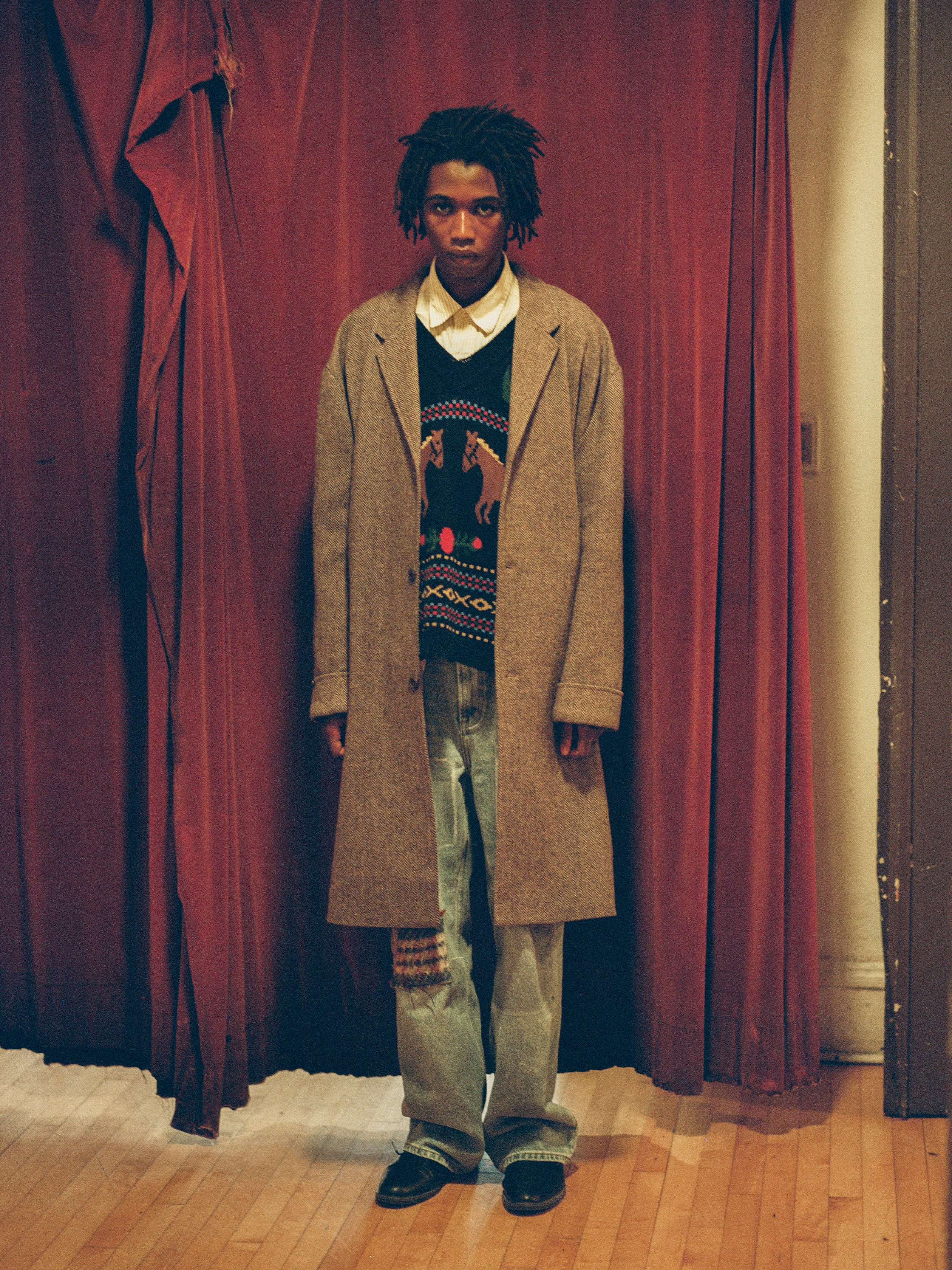 A man sporting a Found Elm Tweed Long Top Coat while standing in front of a red curtain, evoking the timeless history of hunting.