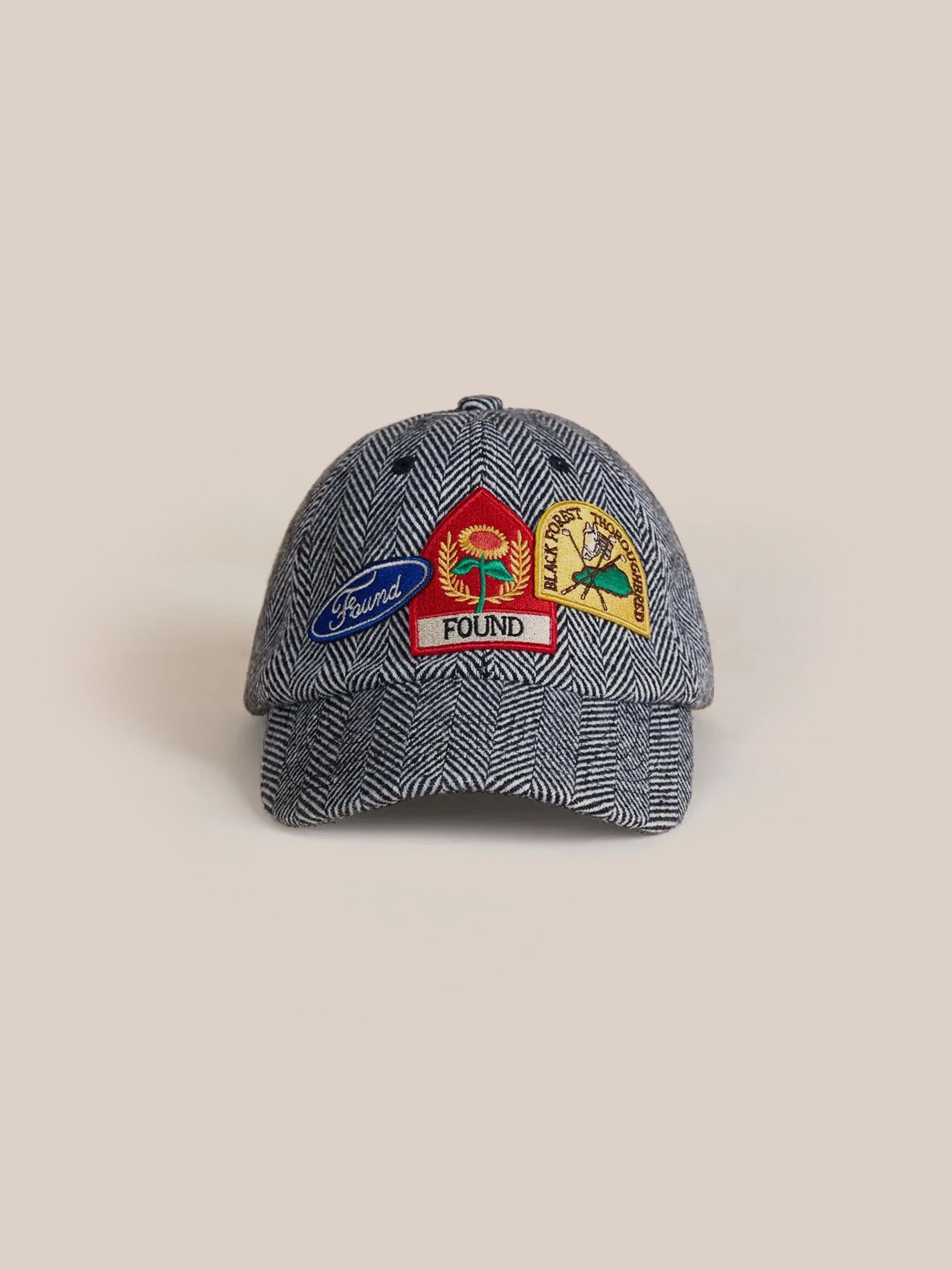 The Herringbone Tweed Patch Cap by Found features a gray design enhanced with vintage patches, including a red and yellow circular emblem, a blue patch labeled "Found," and a yellow patch showcasing a nature image. Ideal for sunny days at the farmers market, it is crafted on a beige base.