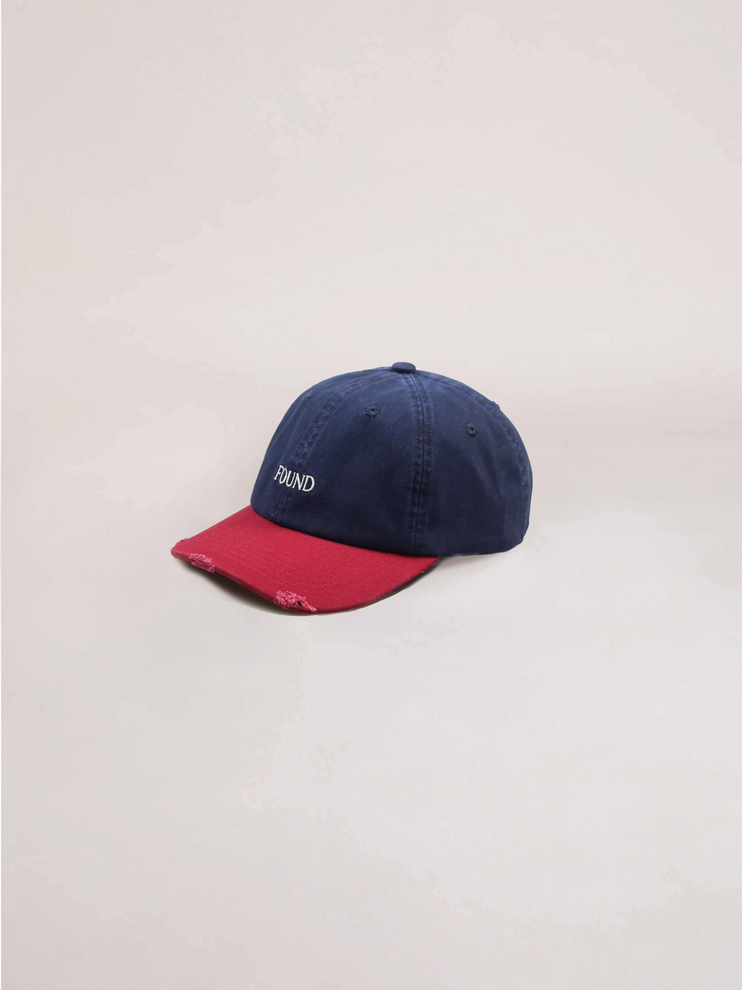 This Two-Tone Logo Cap from the brand FOUND is a navy blue and red cotton cap, showcasing the "FOUND" logo embroidered on the front. It has subtle signs of wear, with slight fraying on the red brim, which enhances its classic character.