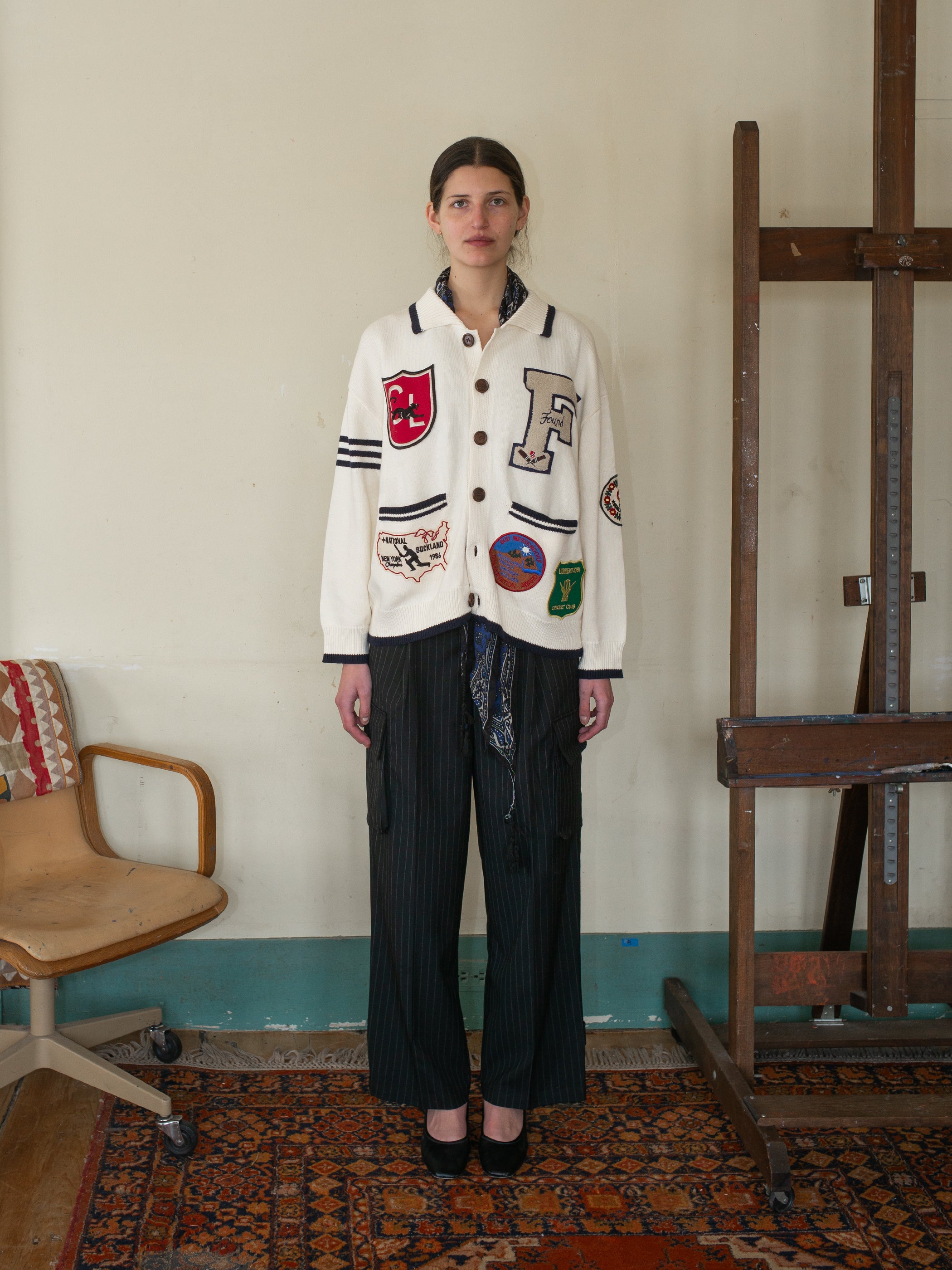 A woman stands in a room exuding collegiate nostalgia, wearing the Found York Varsity Patch Collared Cardigan with striped pants and black heels, beside a wooden easel on a patterned rug.