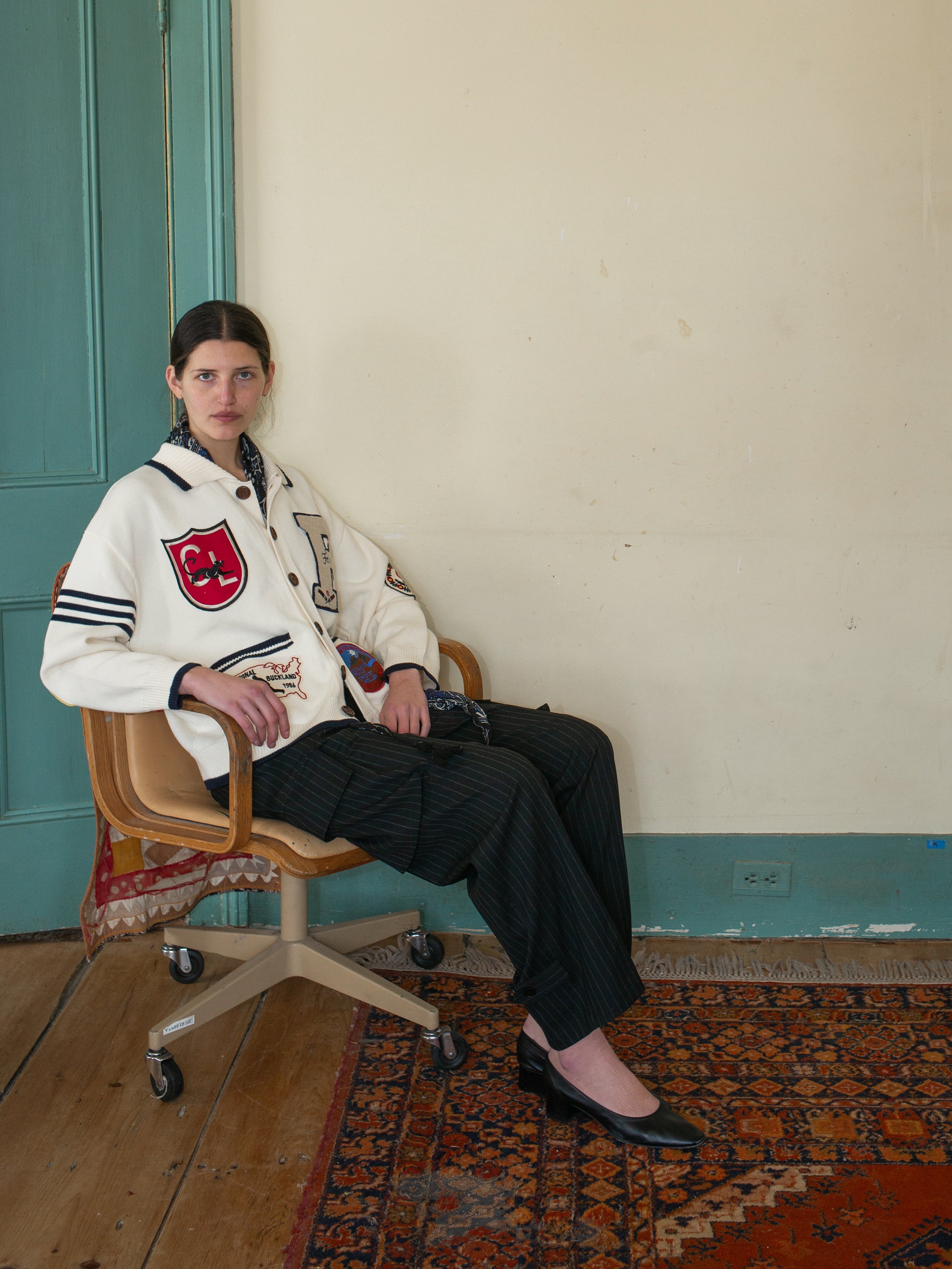 Seated on a swivel chair in a room with light-colored walls and a patterned rug, wearing the Found York Varsity Patch Collared Cardigan, dark pinstripe pants, and black shoes.