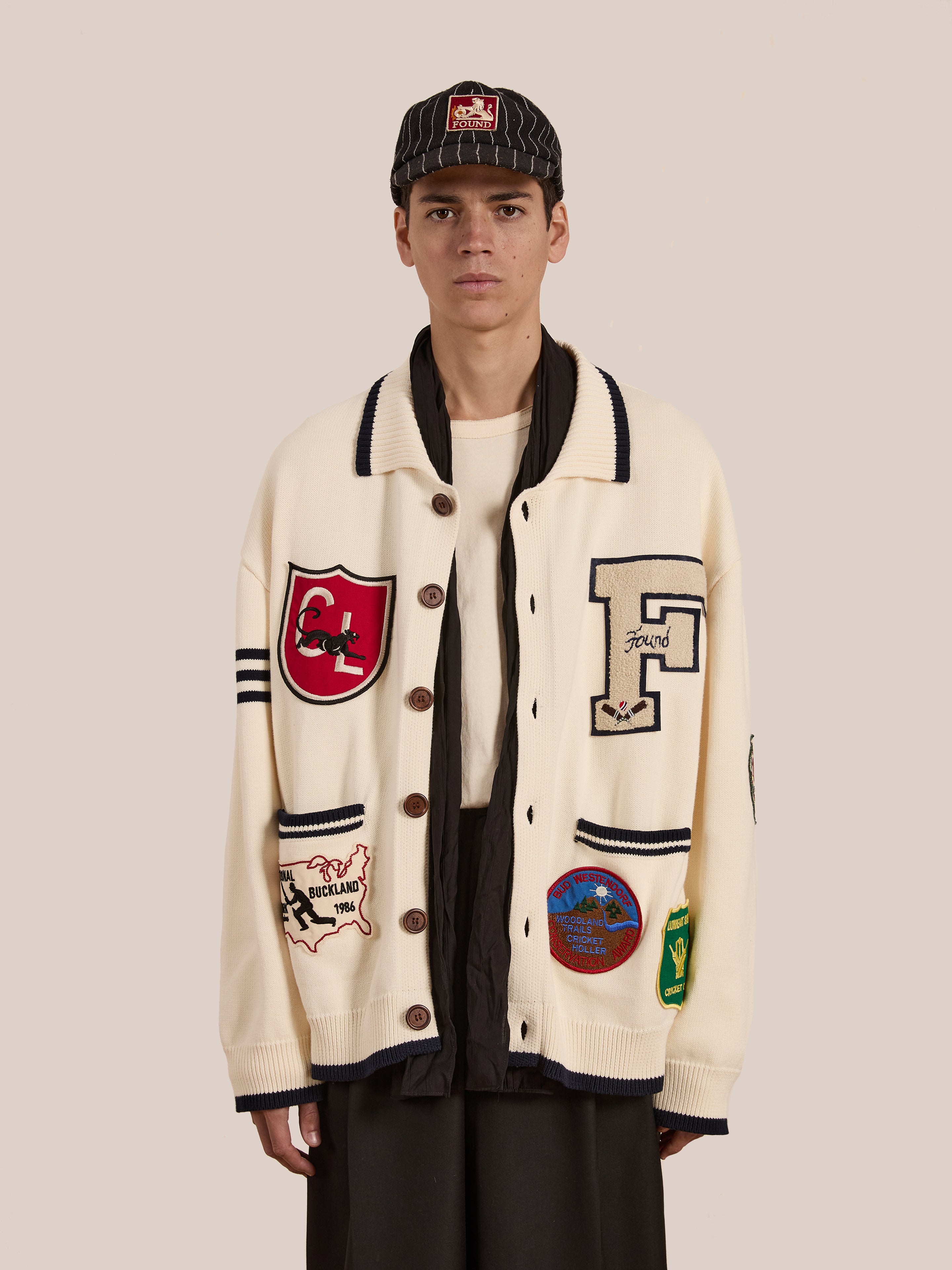 A person wears the FOUND York Varsity Patch Collared Cardigan, exuding collegiate nostalgia with its letters and colorful emblems, over a dark shirt and pairs it with a striped cap to embody a preppy look.