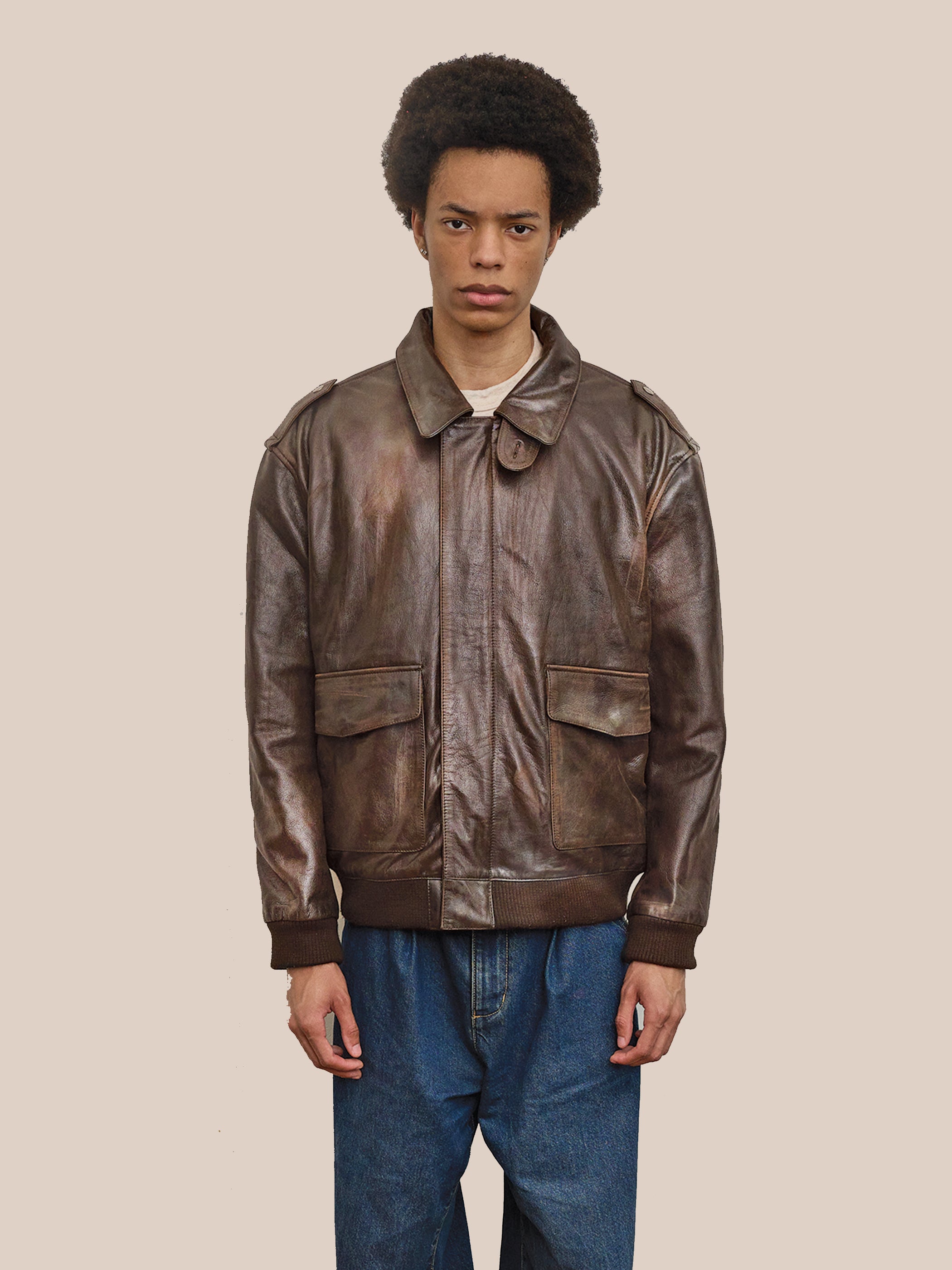 A person wearing the FOUND Distressed Leather Flight Jacket and blue jeans stands against a plain beige background, looking straight ahead with a neutral expression.
