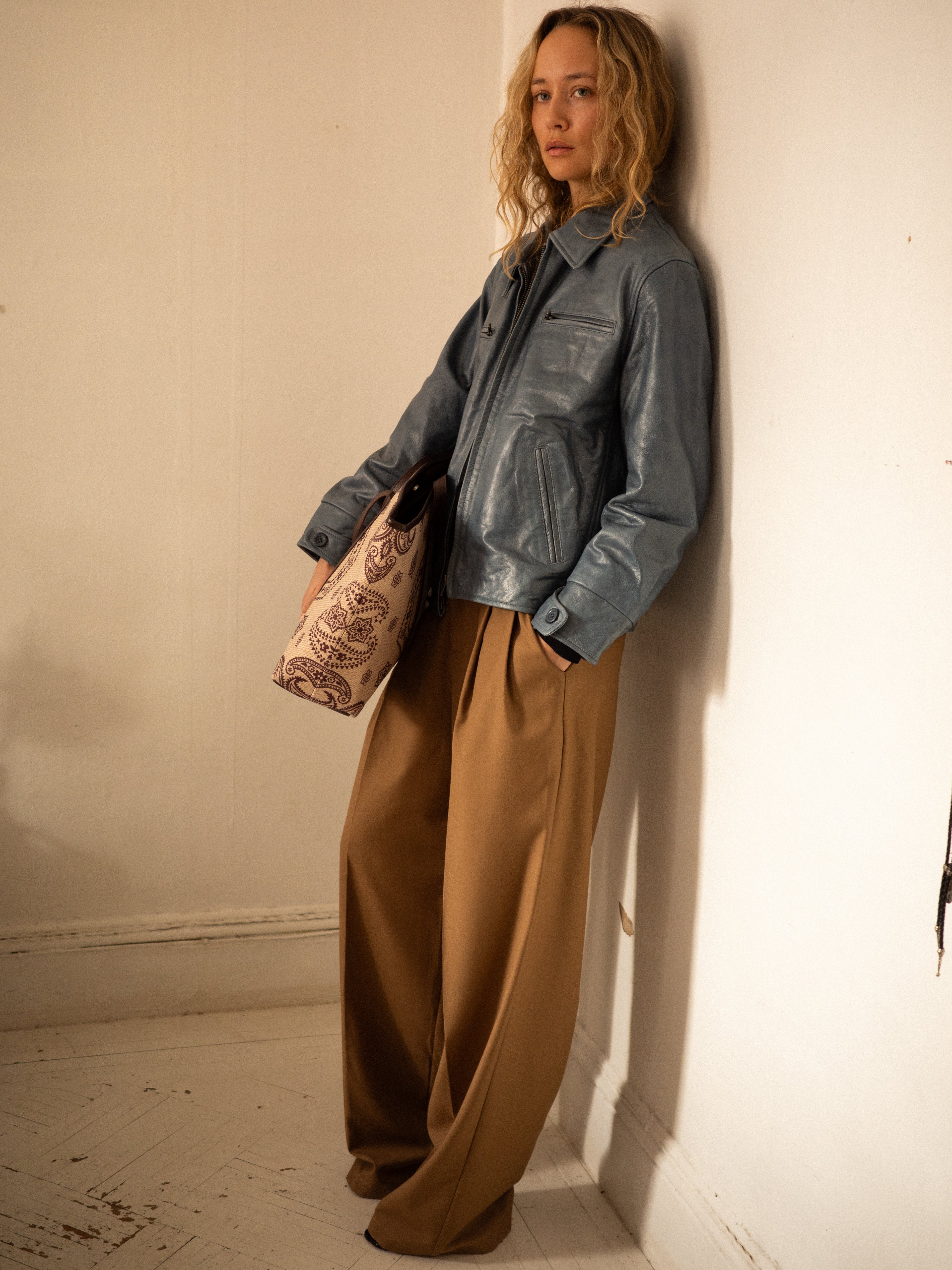 A person with curly hair leans stylishly against a wall, sporting FOUND's Pleated Trousers—a relaxed, high-rise brown option—and a gray leather jacket. A patterned tote bag completes the look.