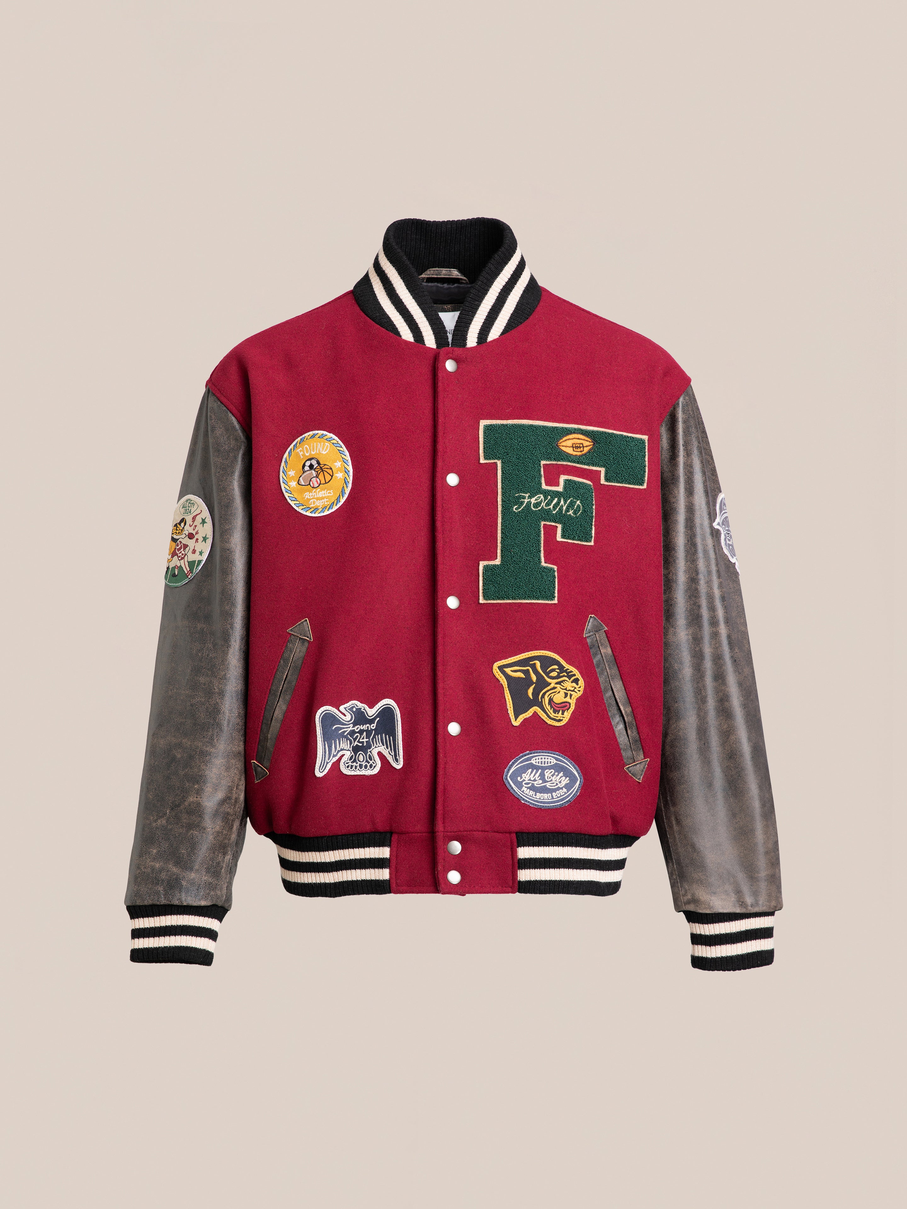 The Found Crimson Letterman Jacket presents a vintage appeal with its red body, black and white striped trims, and dark brown leather sleeves. Made from ethically sourced materials, it showcases various patches including a prominent green and yellow "F" along with multiple logos on the front and sleeves.