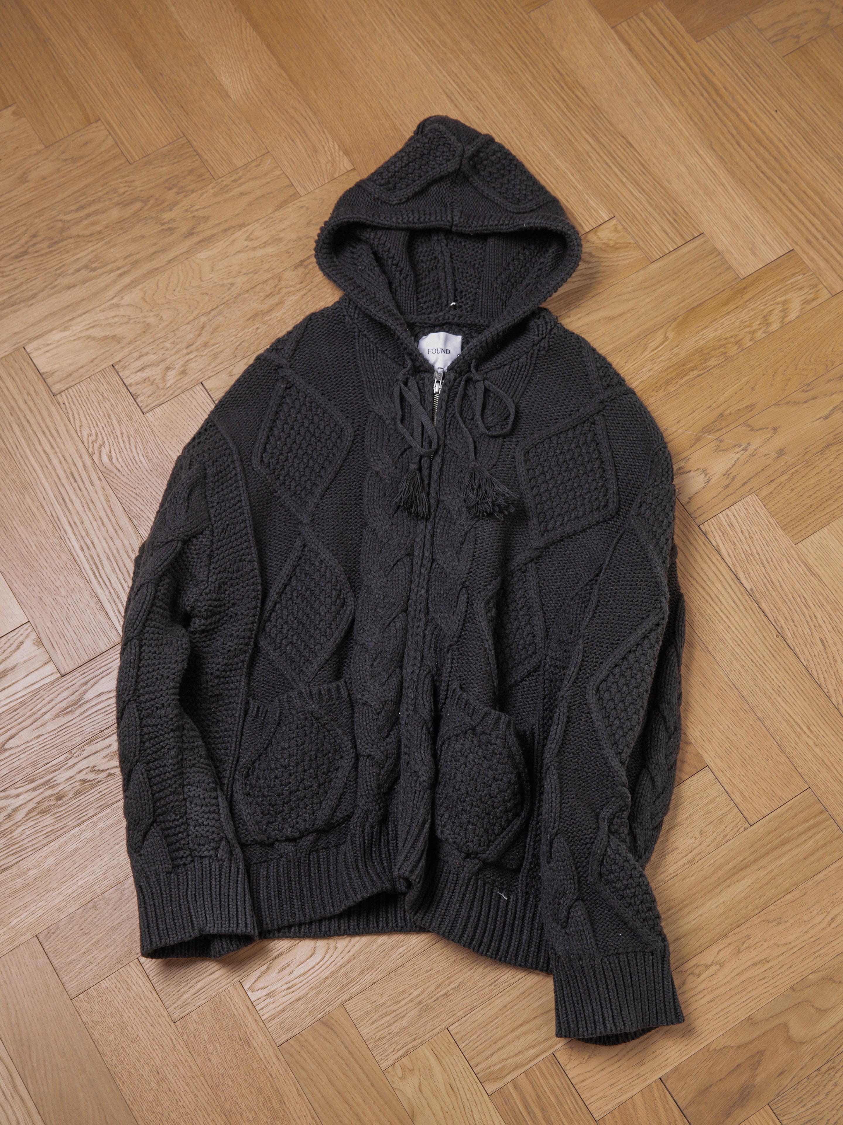 A dark gray, chunky knit **Found Zip Up Cable Knit Hoodie** with a front zipper and two pockets, laid flat on a wooden herringbone-pattern floor, offers comfort and warmth.